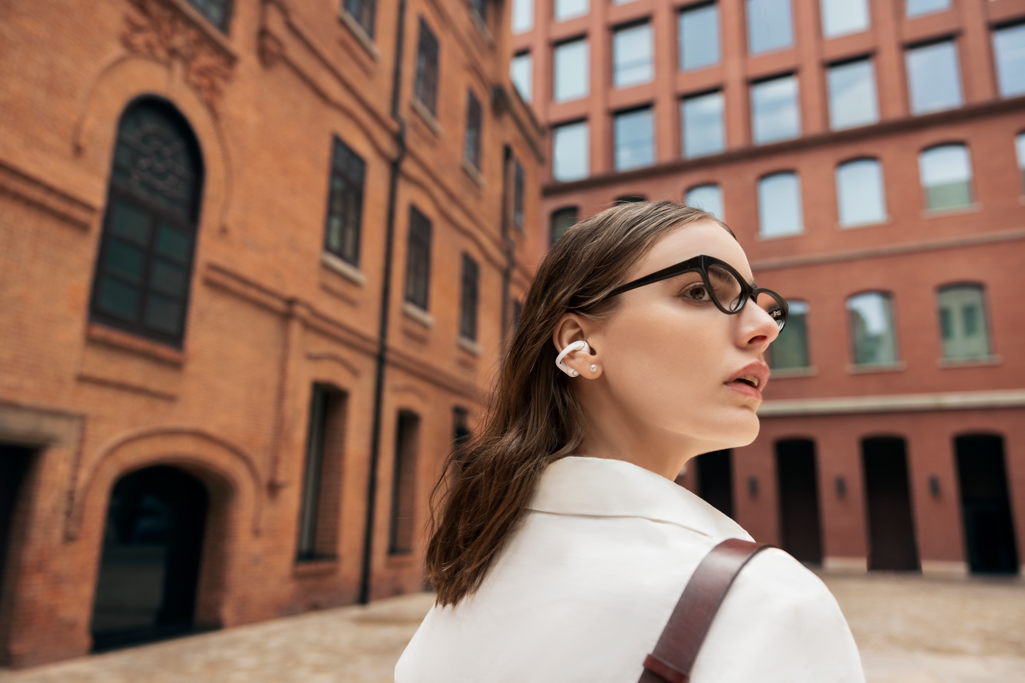 Frau mit Brille und Ohrhörern steht vor Backsteingebäuden.