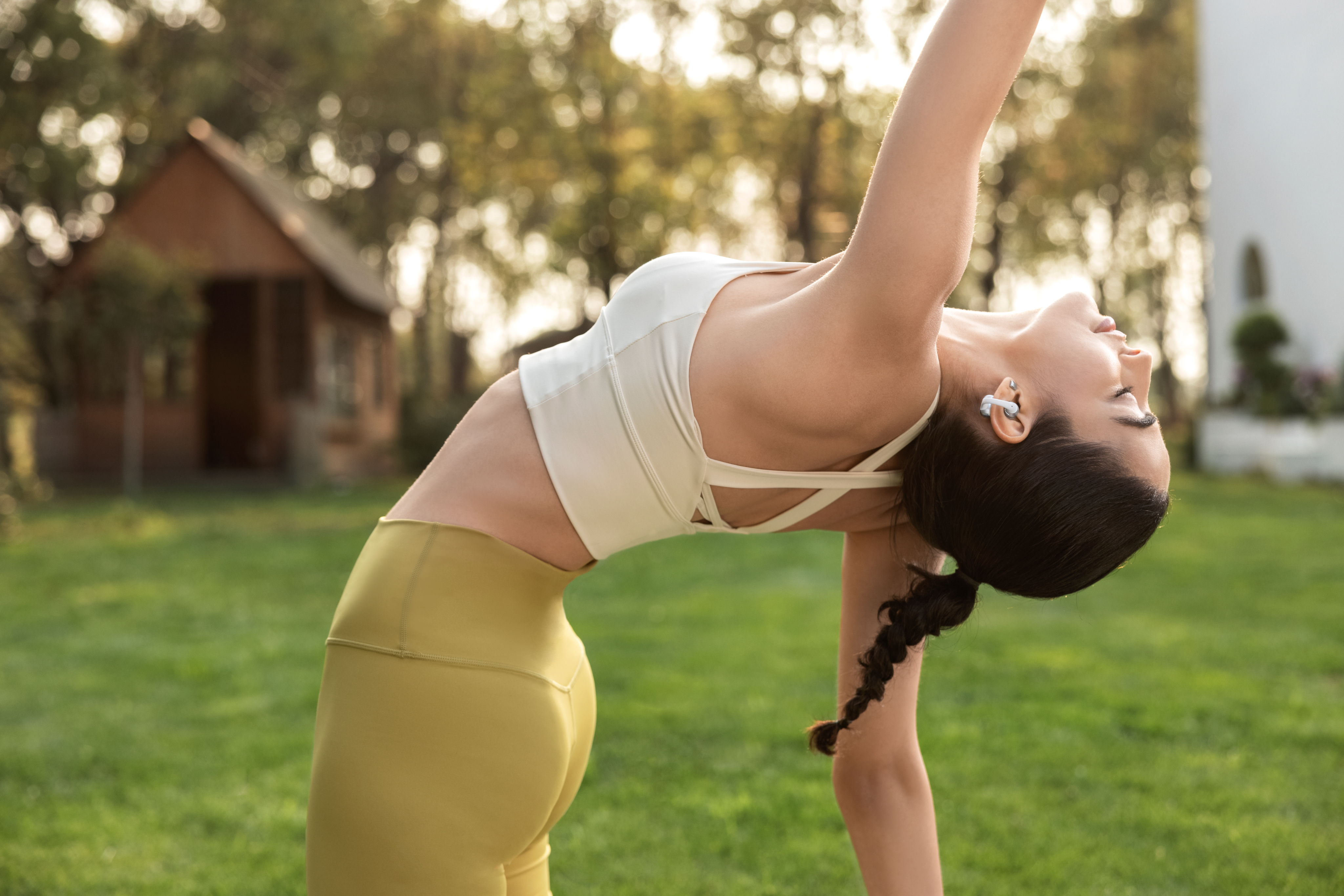 Frau in gelber Hose und weißem Top macht Yoga im Freien, mit einem Haus im Hintergrund.