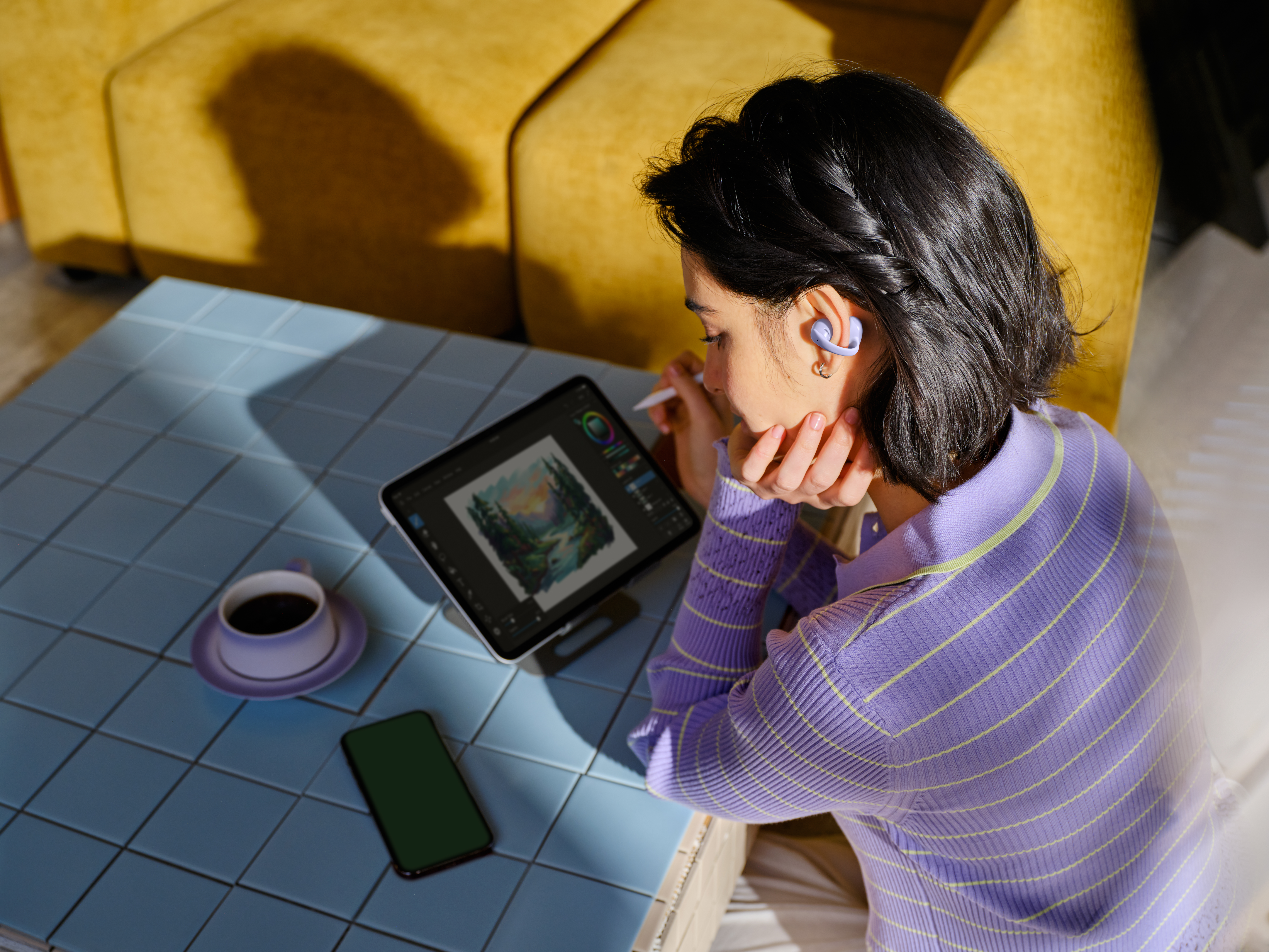 Eine Frau mit violetten Ohrhörern zeichnet auf einem Tablet. Kaffee und Handy liegen auf dem Tisch.
