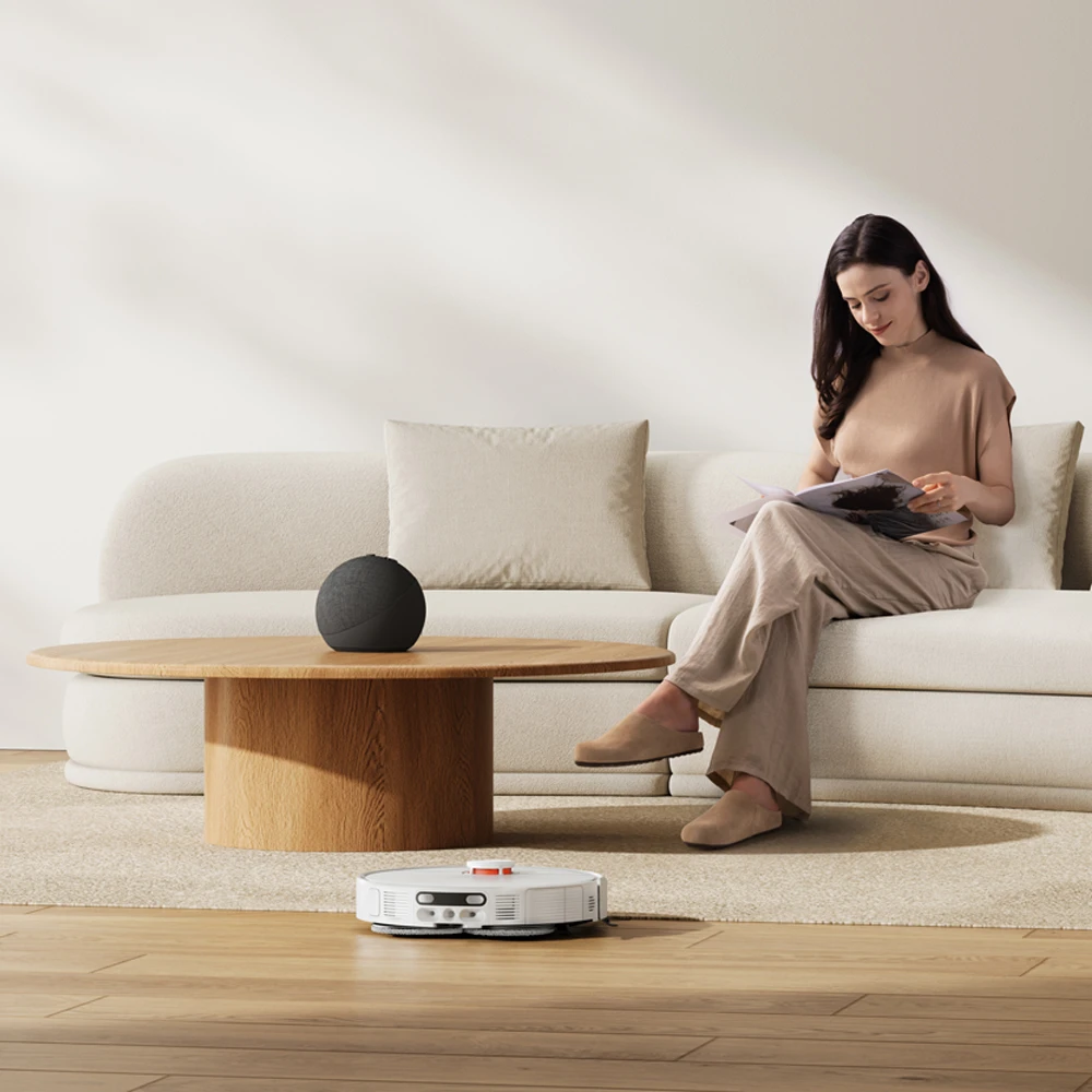 Une femme lit sur un canapé pendant qu'un robot aspirateur nettoie le sol.