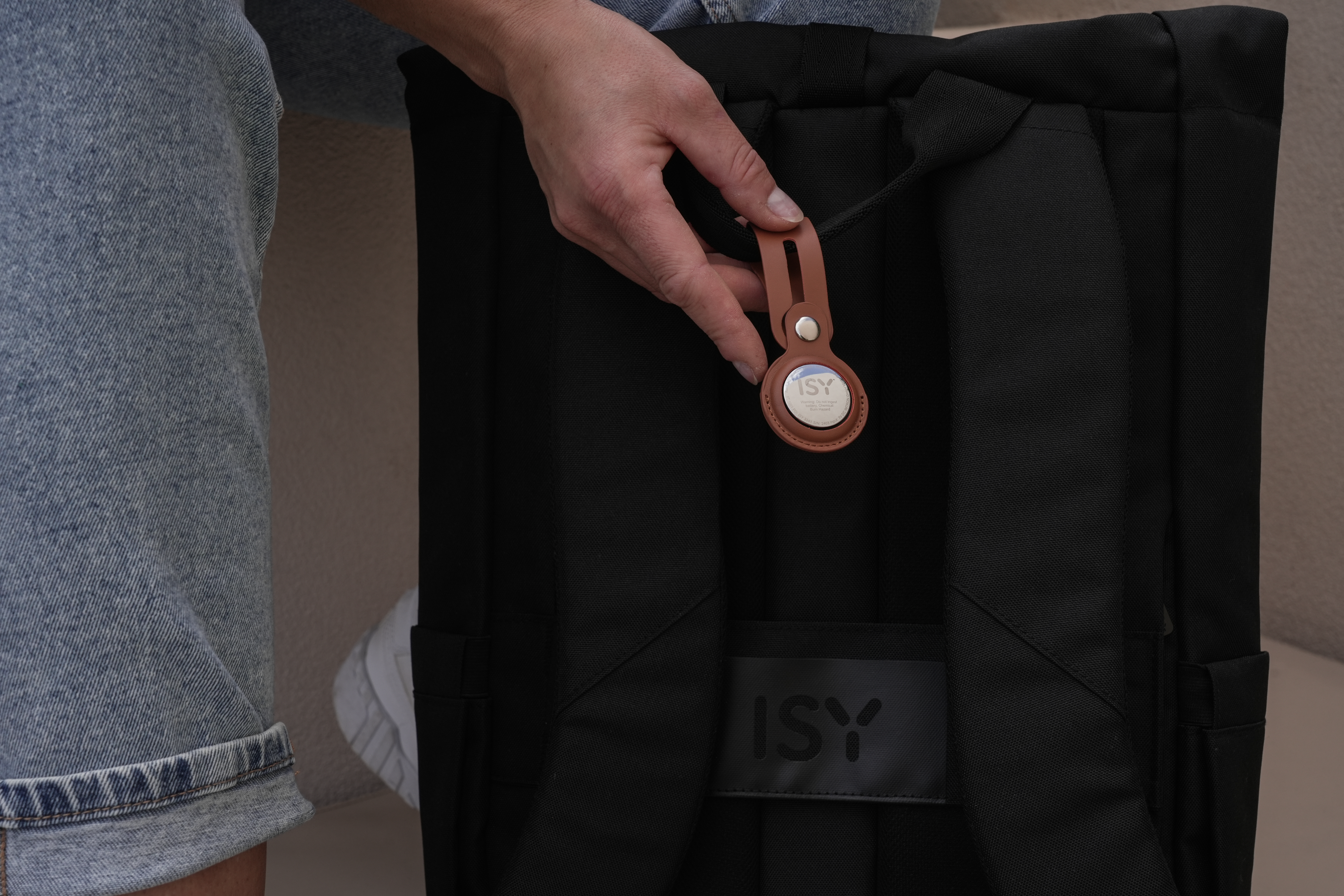 Eine Hand befestigt einen braunen Leder-Trackerhalter an einem schwarzen Rucksack.