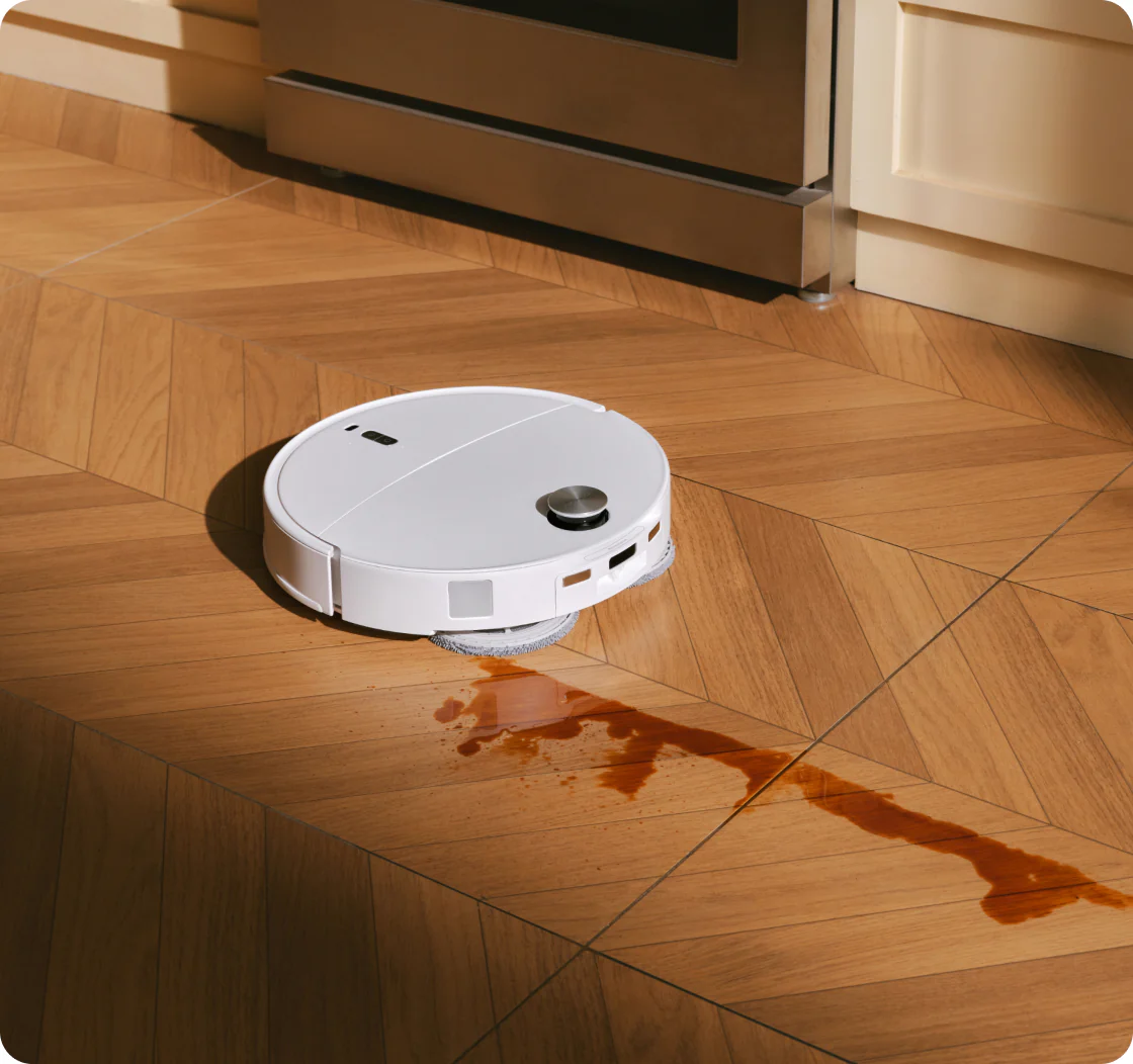 Un robot aspirapolvere bianco pulisce un pavimento in legno con liquido marrone versato.