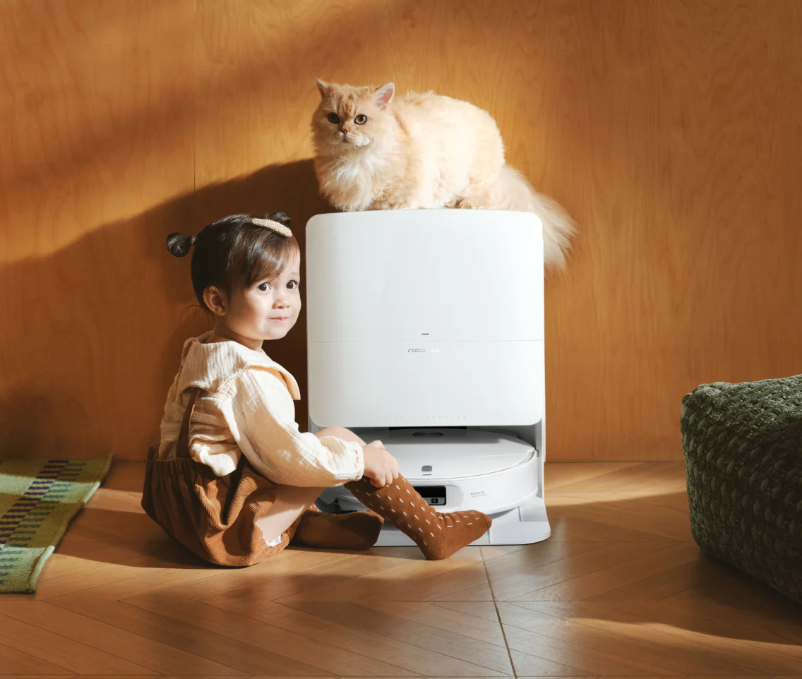 Una bambina e un gatto siedono vicino a un robot aspirapolvere bianco su un pavimento di legno.