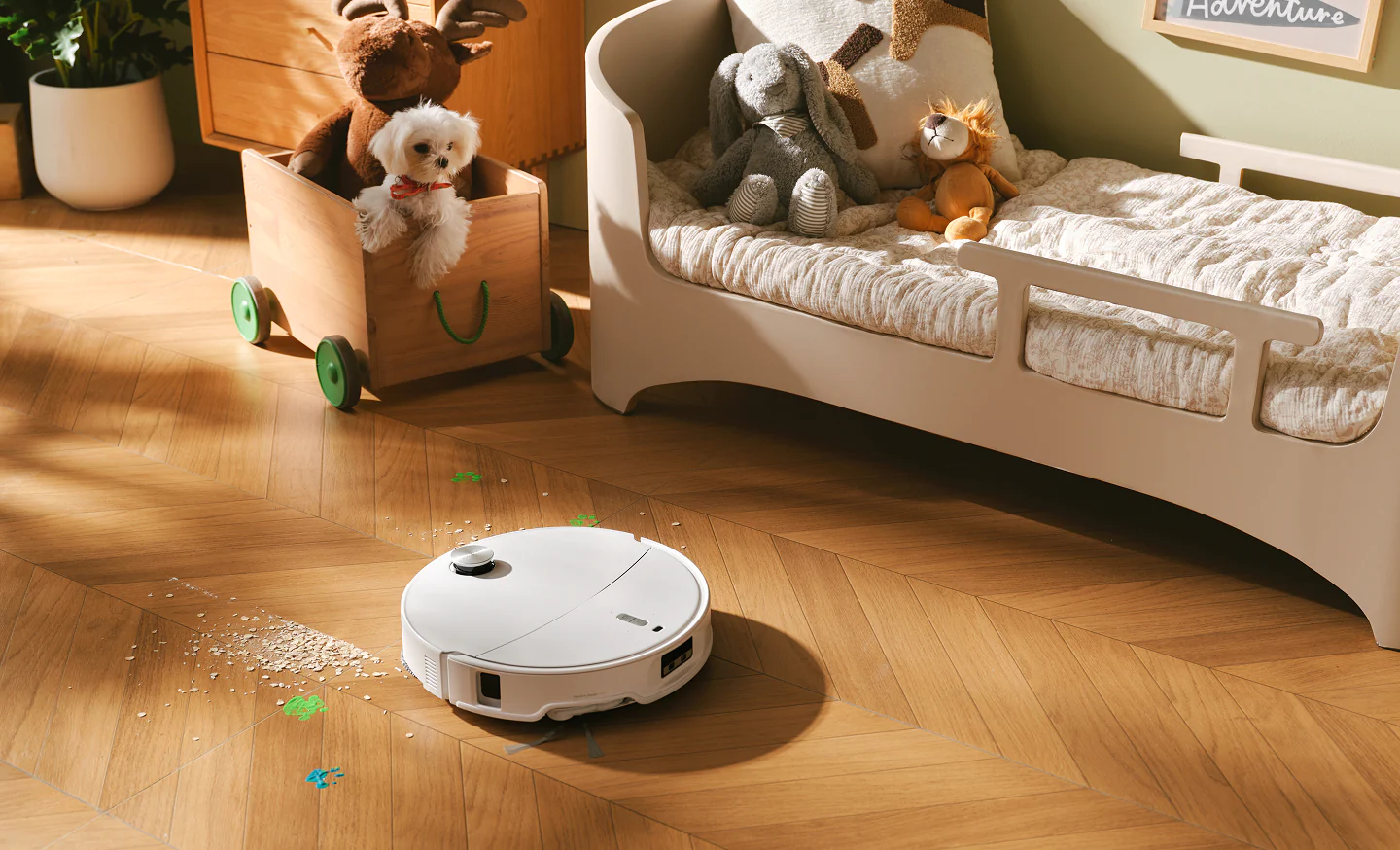 Un robot aspirapolvere bianco pulisce un pavimento in legno vicino a un lettino con peluche.