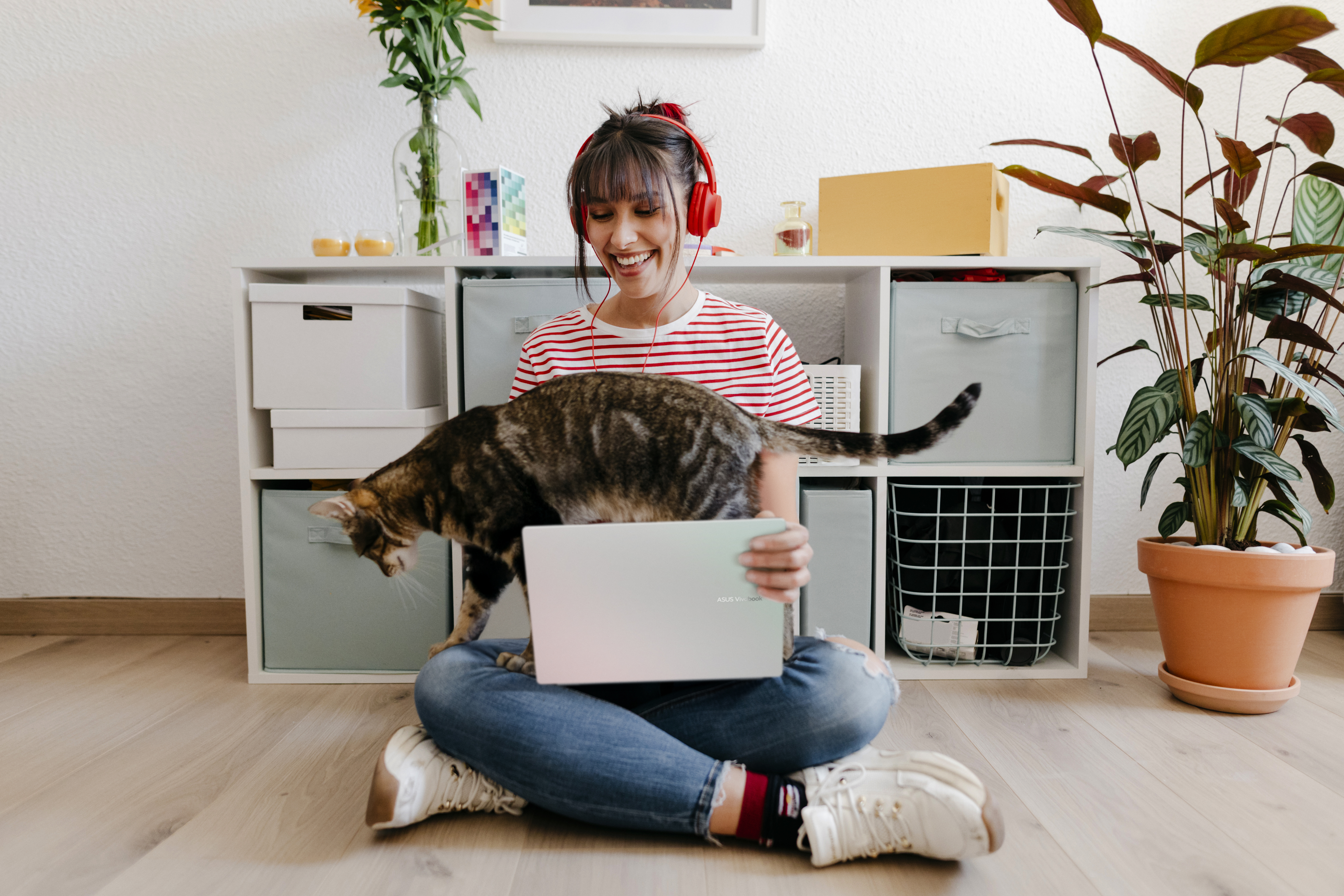 Een vrouw met koptelefoon zit op de grond, kijkend naar een laptop, met een kat op haar schoot.