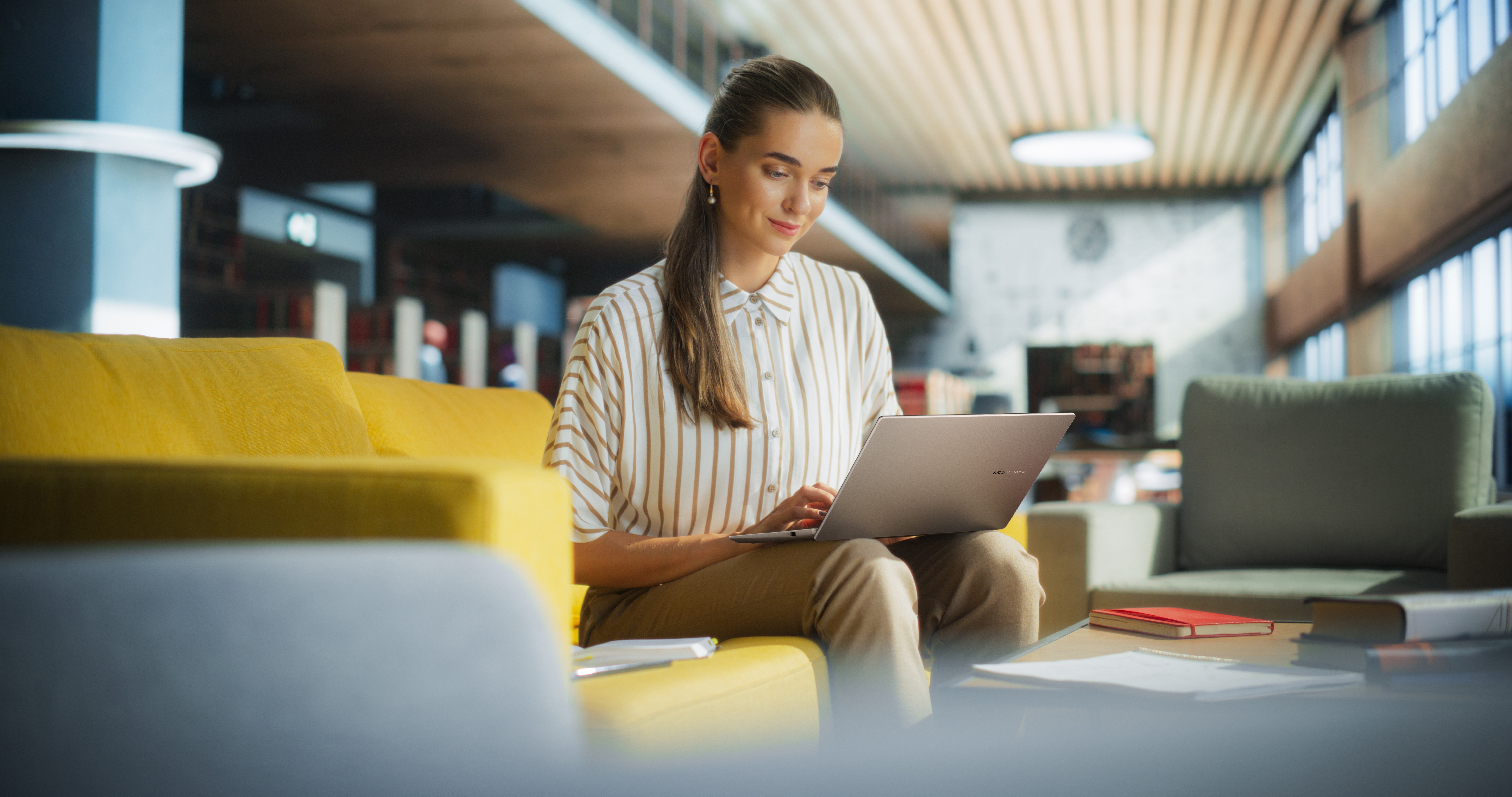 Vrouw werkt op laptop op gele bank in modern kantoor.