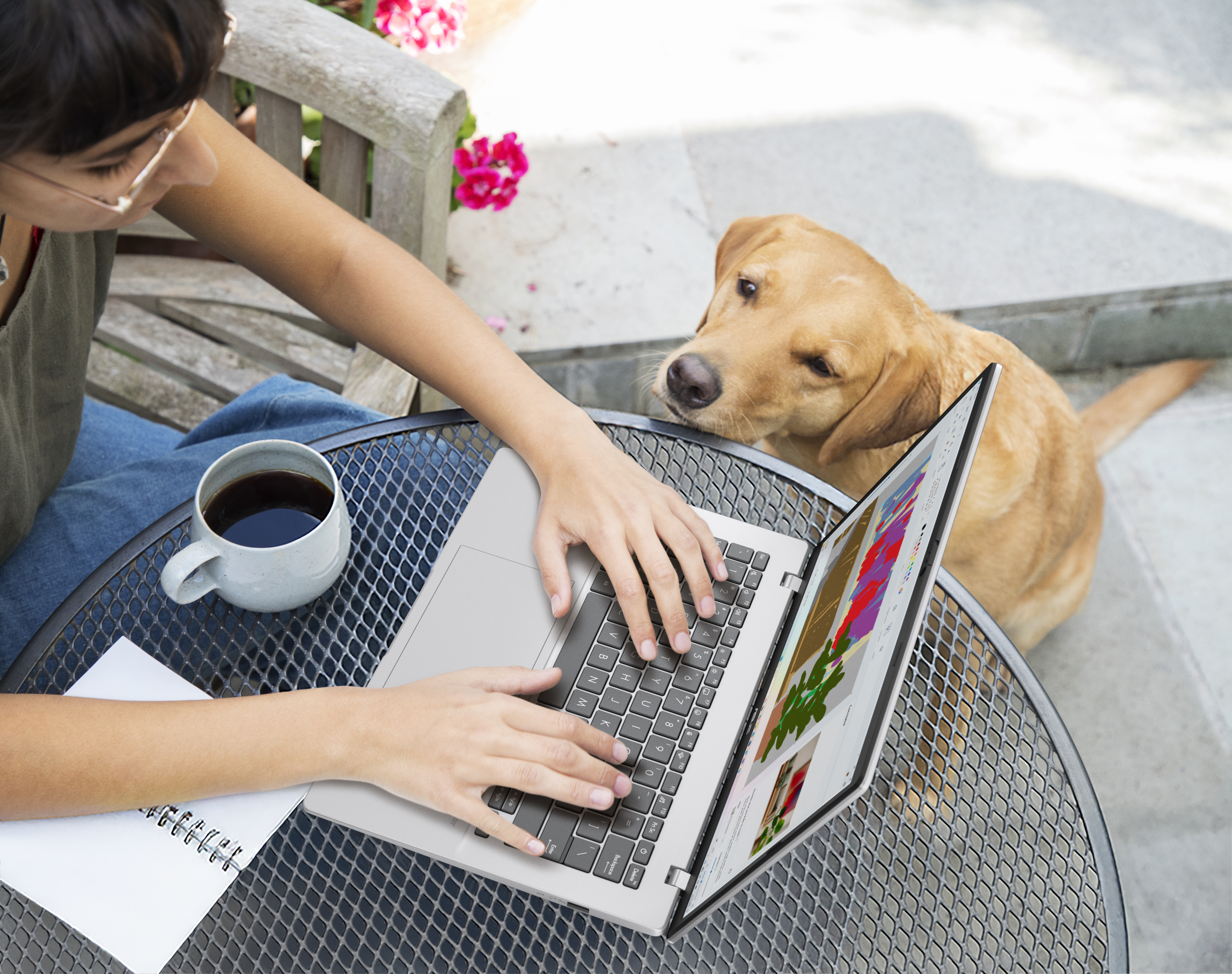 Vrouw typt buiten op laptop met hond die toekijkt. Koffie en notitieboekje in de buurt.