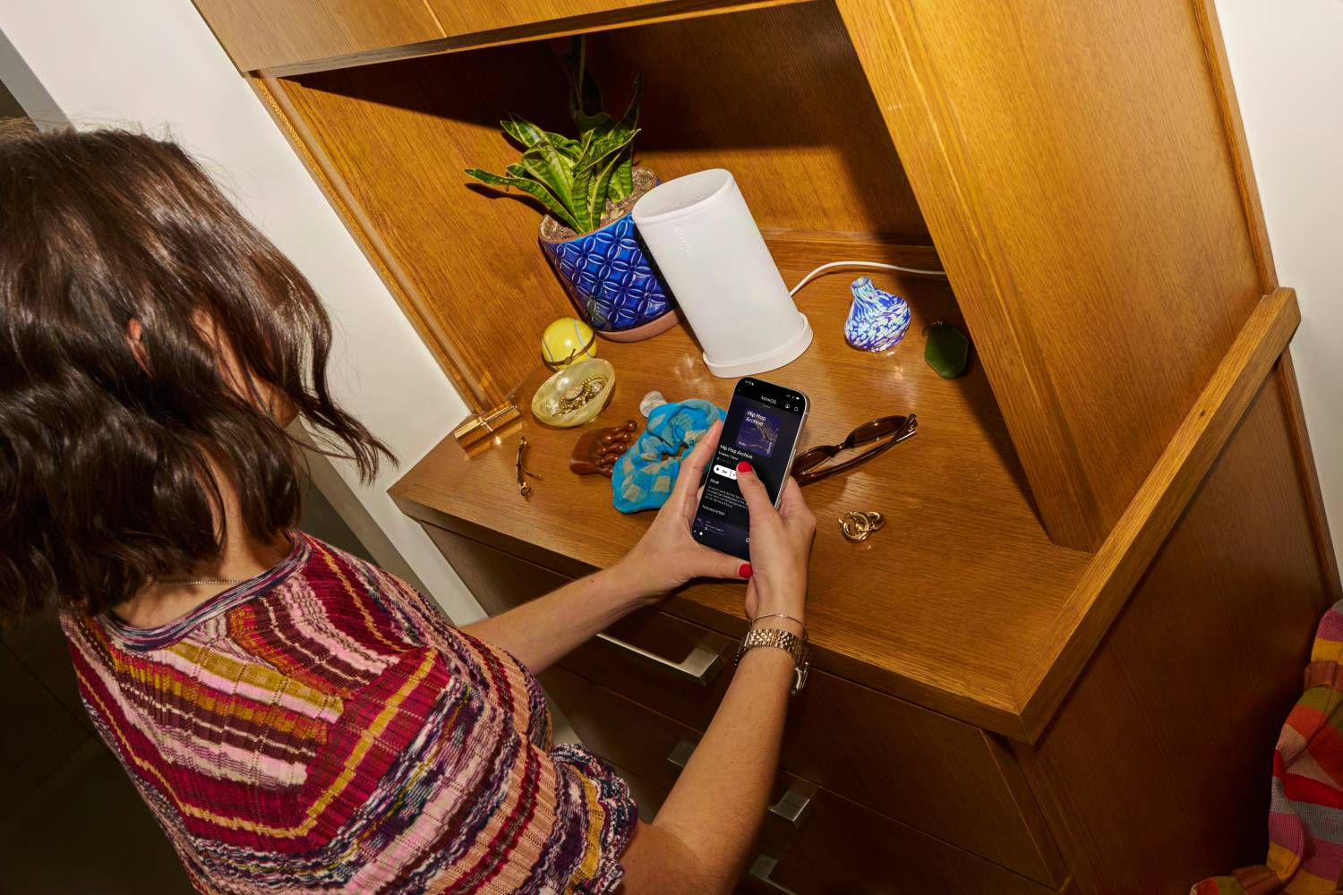 Mujer con teléfono frente a estante de madera con decoración, planta, altavoz.