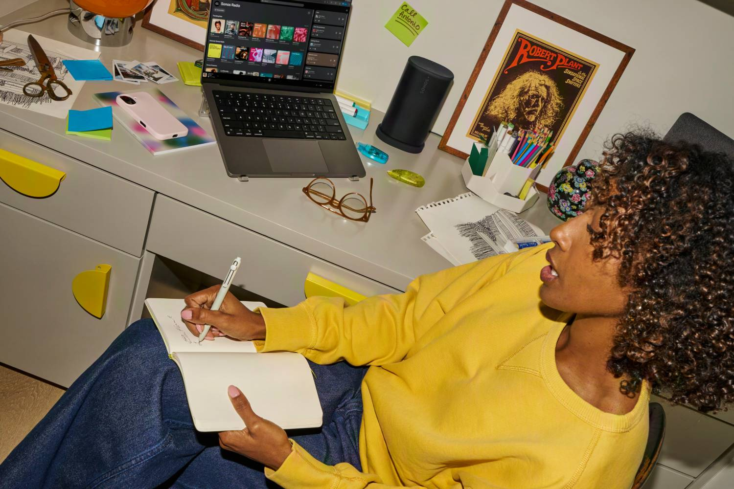 Mujer escribiendo en cuaderno en escritorio con laptop, altavoz, póster.