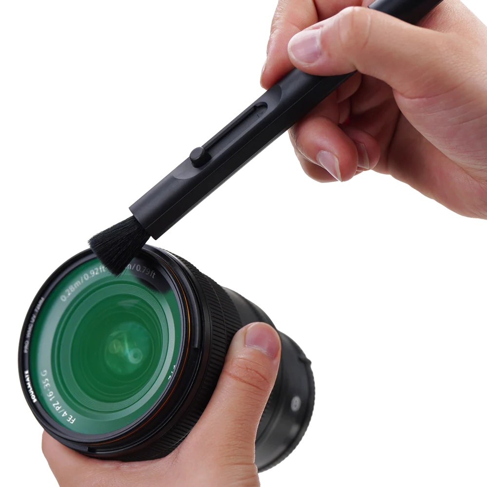 Una mano tiene un obiettivo della fotocamera e un'altra mano usa uno strumento di pulizia nero per spazzolare l'obiettivo.