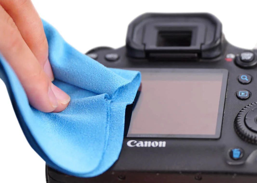 Una mano pulisce lo schermo di una fotocamera Canon nera con un panno in microfibra blu.