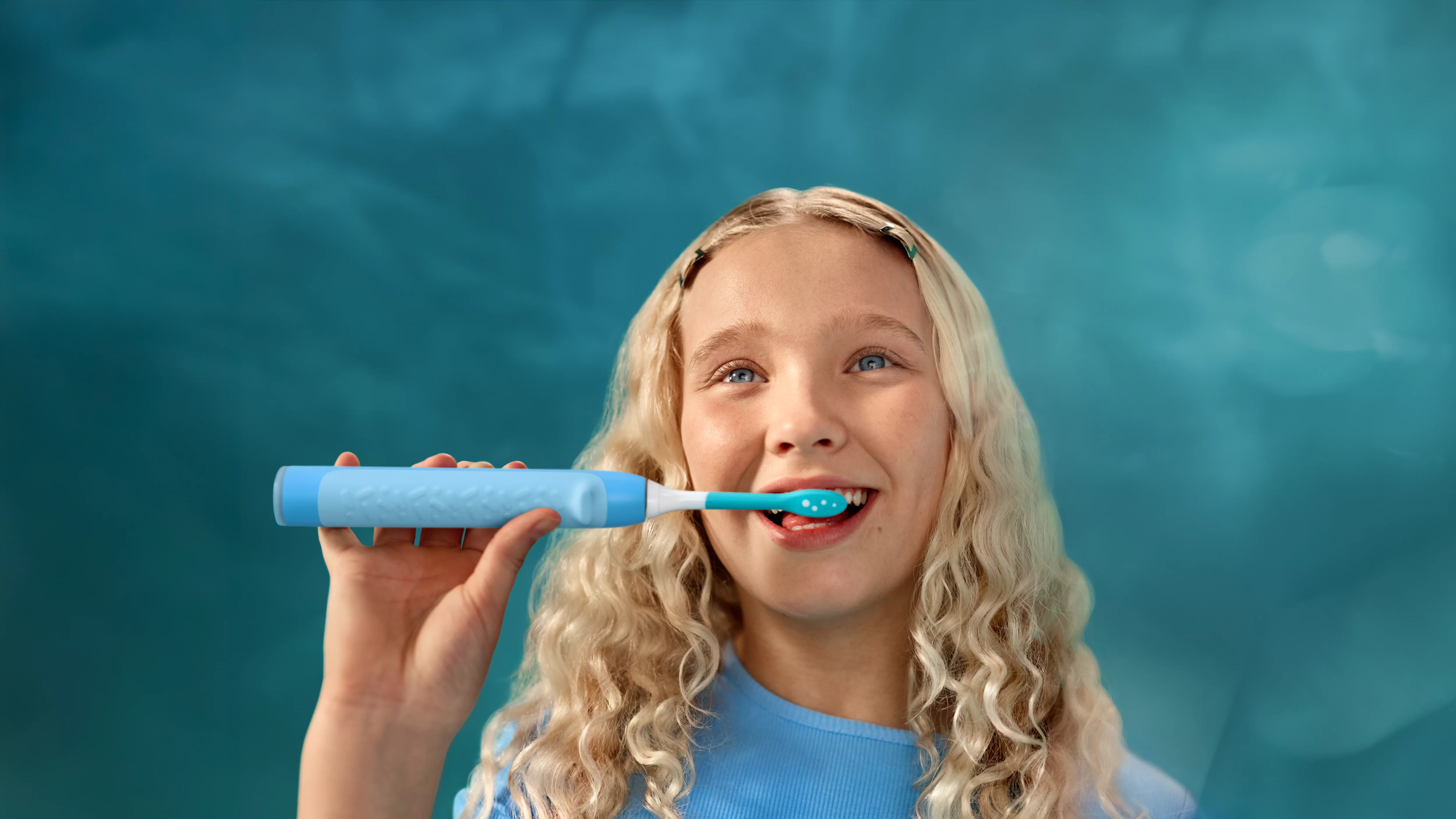 Una ragazza bionda si lava i denti con uno spazzolino elettrico blu.