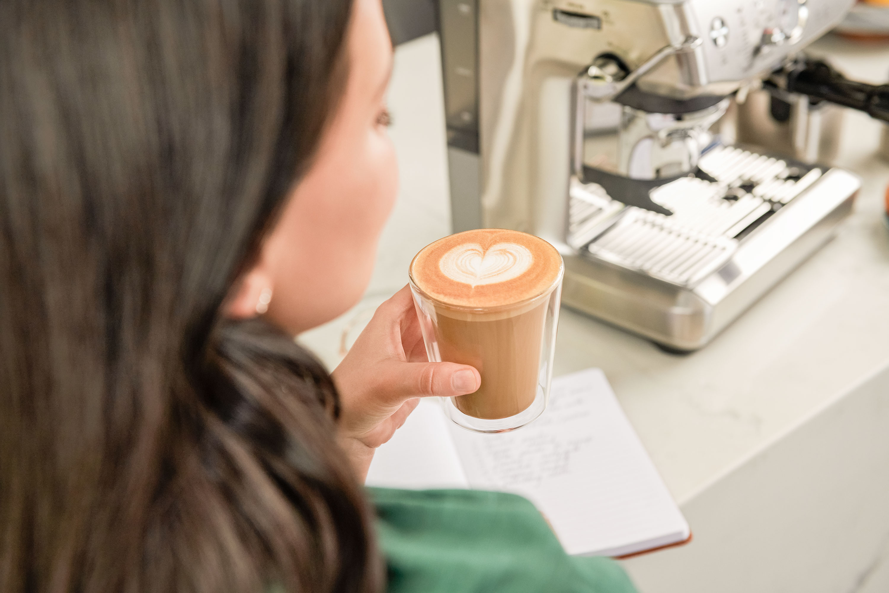Nő pohárban tartott latte art kávét, kávégép és jegyzetfüzet mellett.