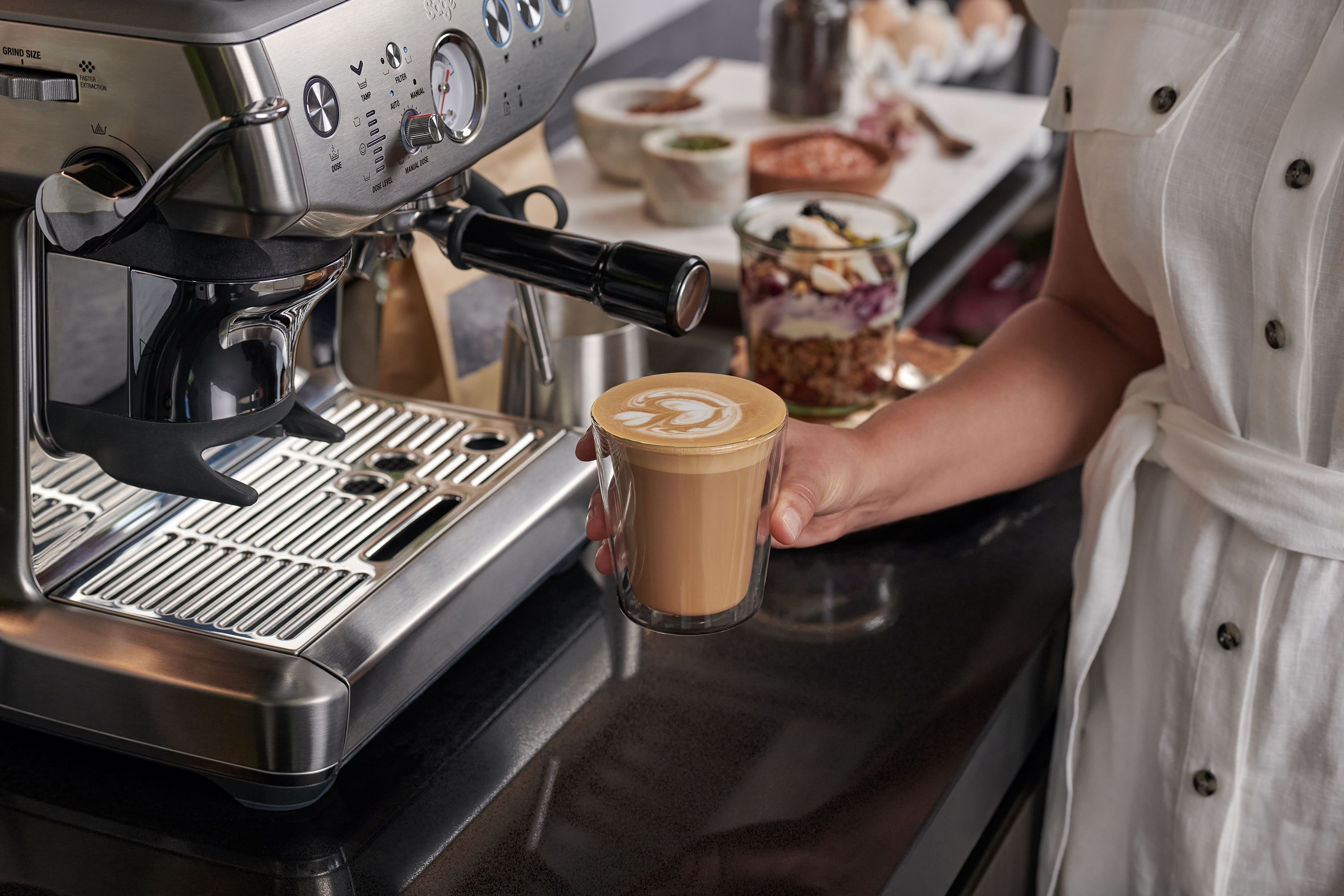 Egy kéz egy pohár lattét tart egy eszpresszógép mellett.