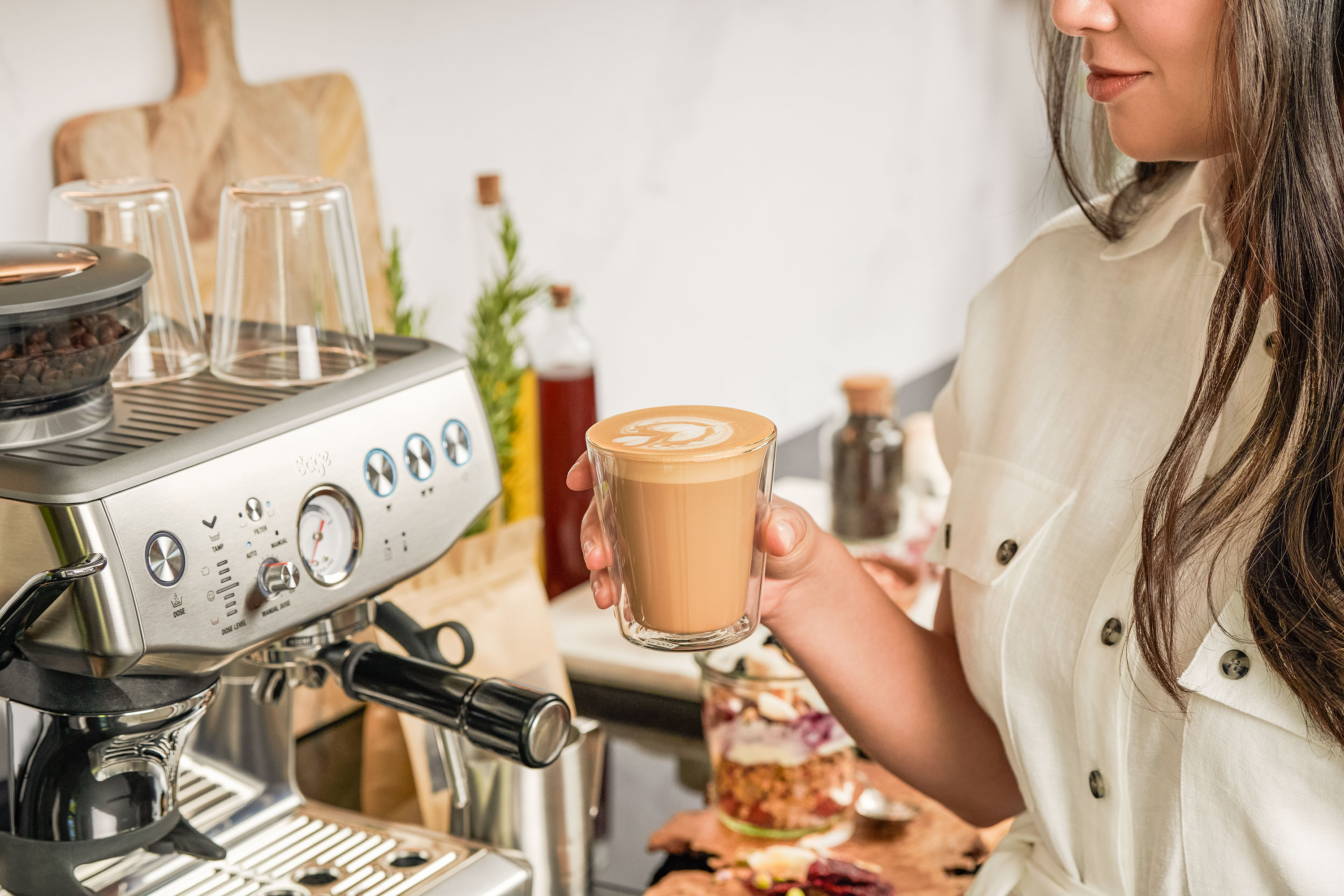 Egy nő egy pohár latte art kávét tart egy eszpresszógép mellett.