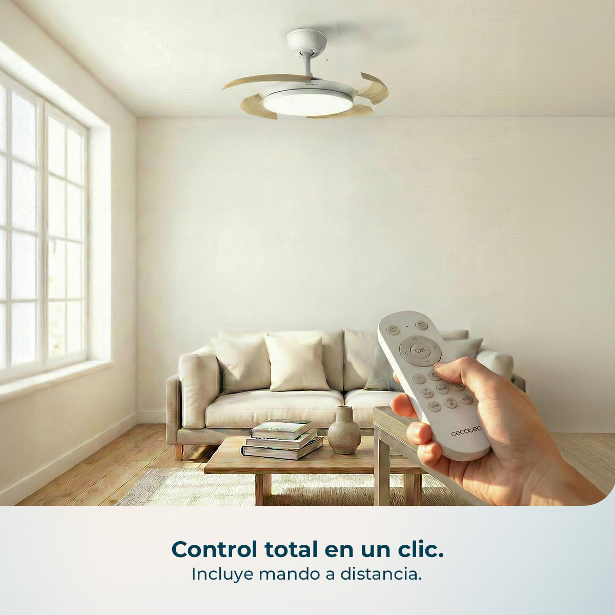 Una mano sostiene un mando a distancia frente a una sala con ventilador de techo y sofá.