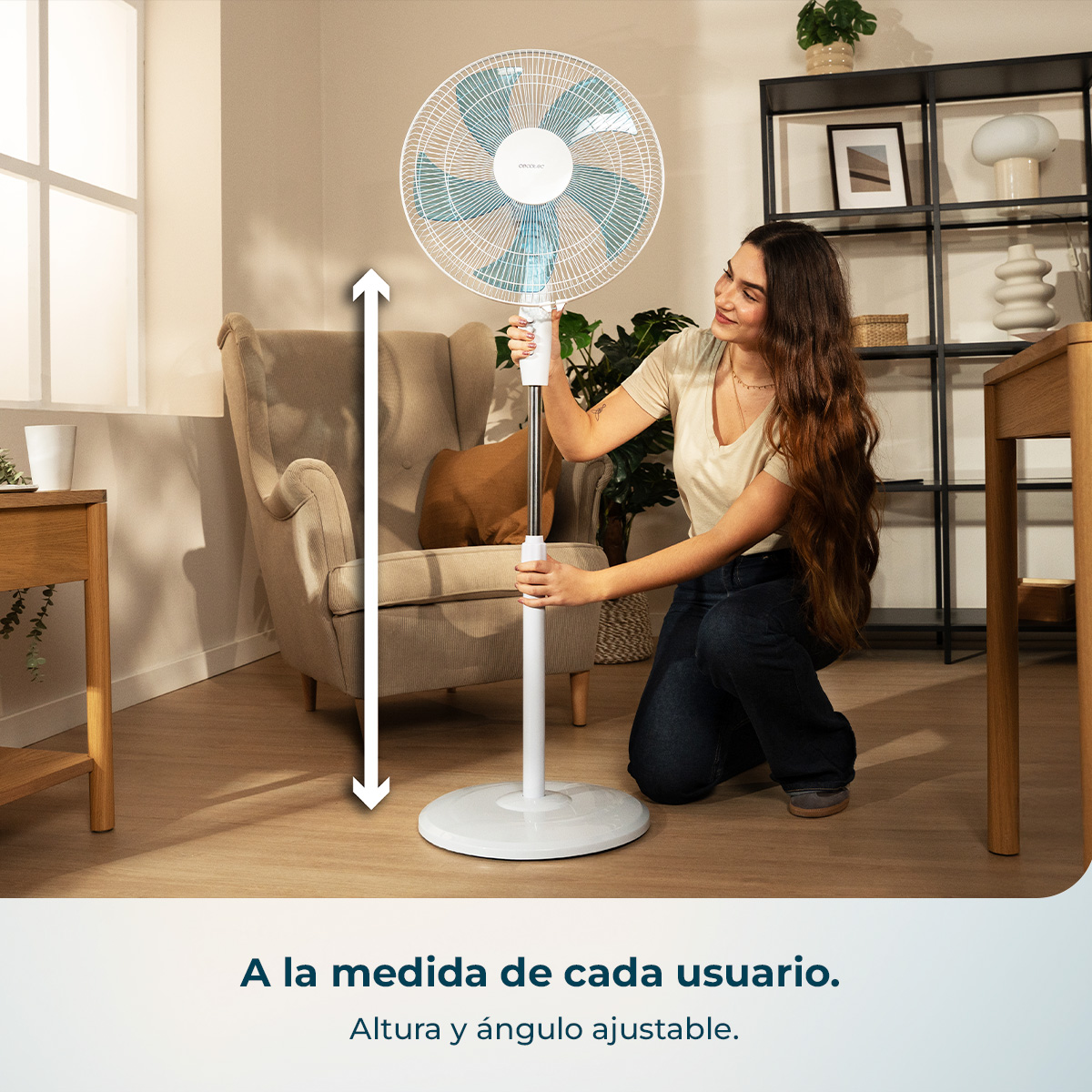 Una mujer ajusta la altura de un ventilador de pie blanco. La habitación está iluminada.