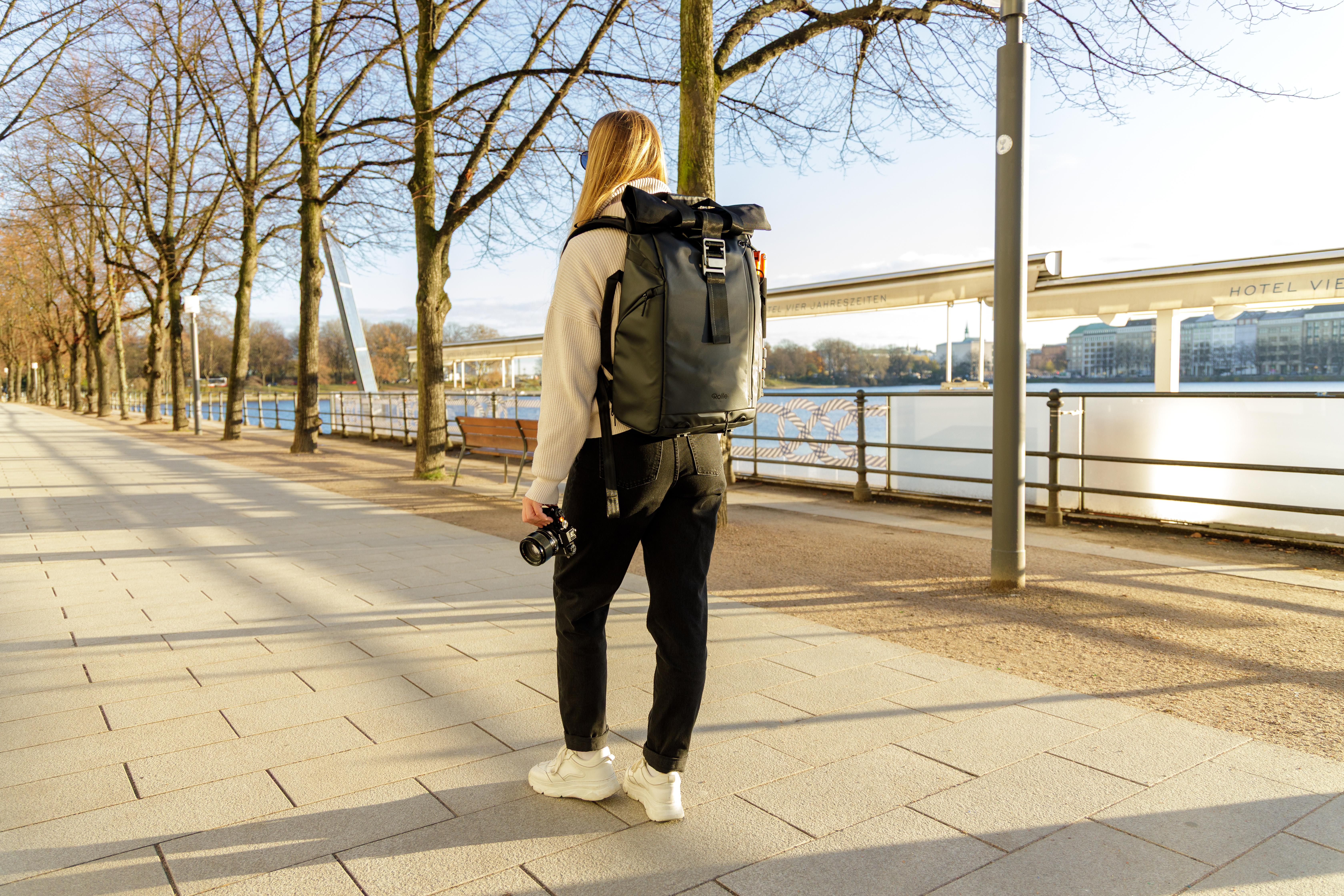 Frau mit Rucksack und Kamera geht auf einem Gehweg am Wasser entlang.