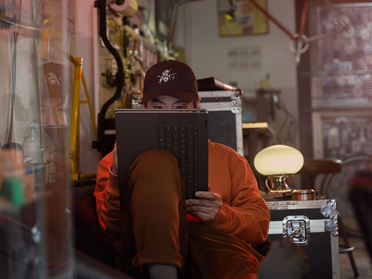 A man wearing a cap sits using a laptop. He wears an orange sweatshirt.