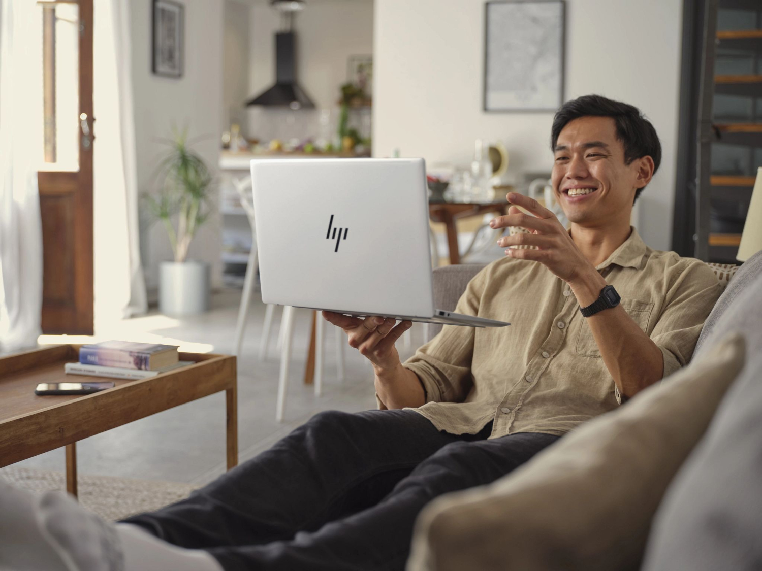 A smiling man holds a laptop on a couch, gesturing with his hands. A coffee table is nearby.