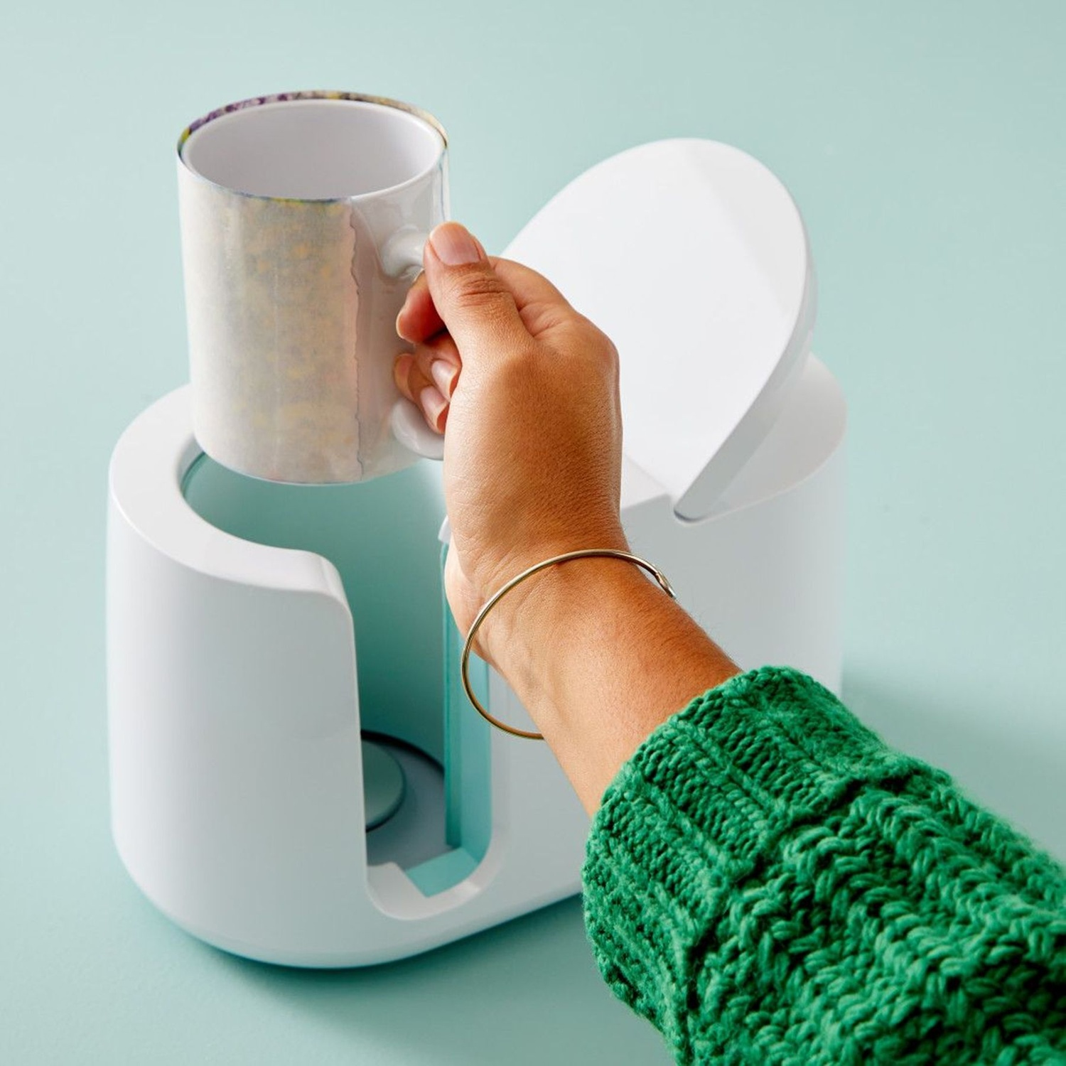 Una mano inserisce una tazza fantasia in una macchina per sublimazione bianca su sfondo verde acqua.