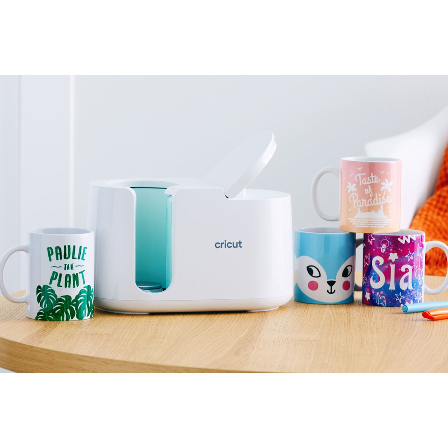 Una pressa per tazze Cricut bianca con quattro tazze decorate davanti su un tavolo di legno.