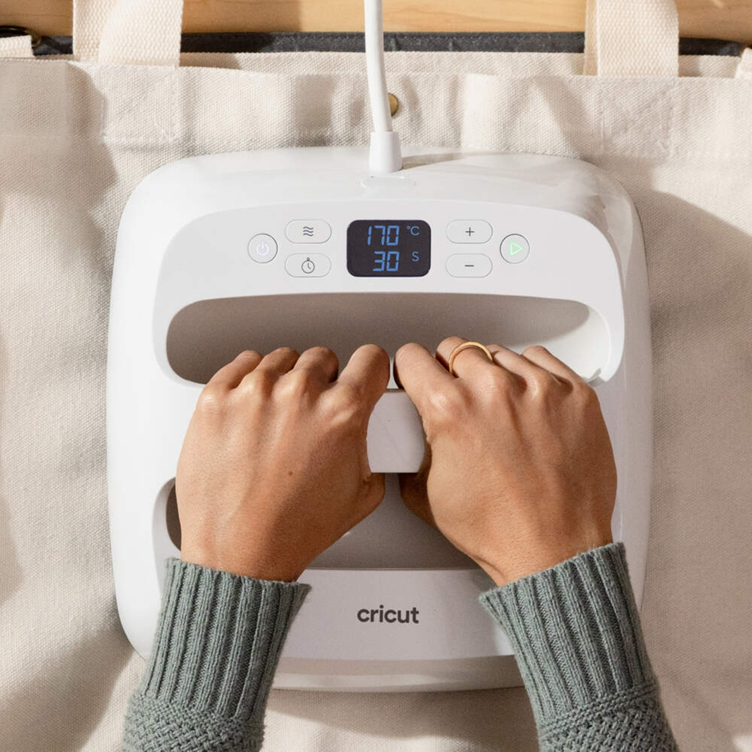 Mani azionano una pressa Cricut bianca su tessuto con display digitale.