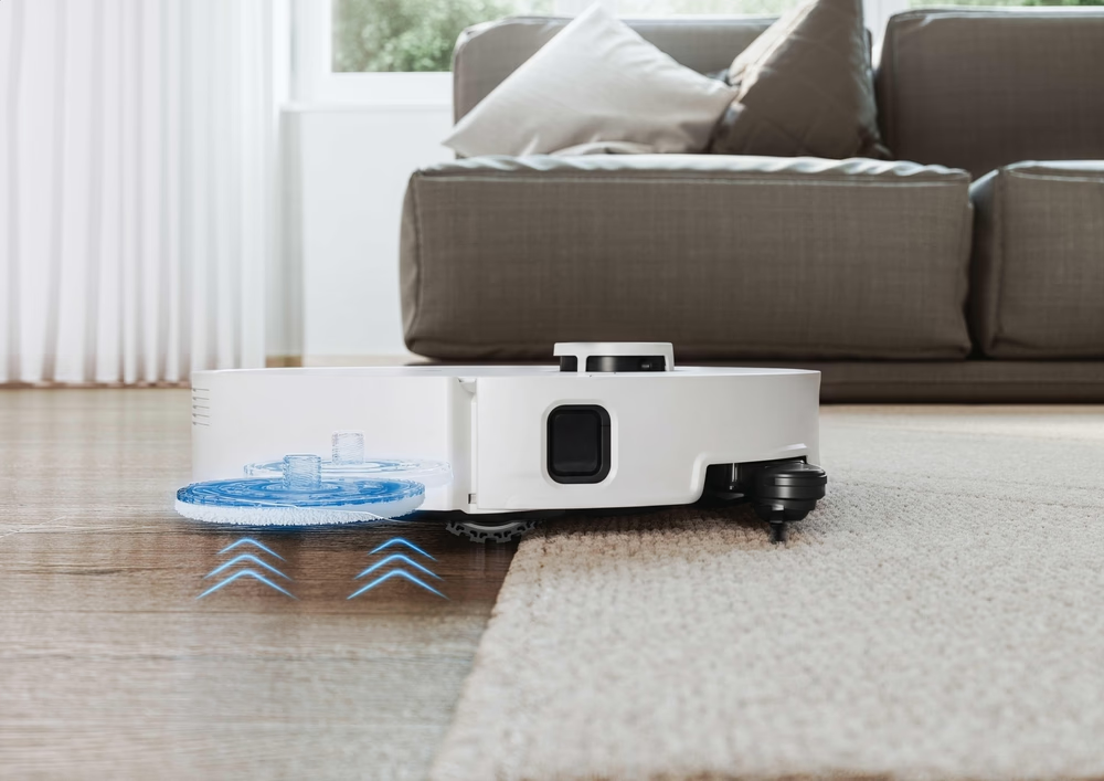 Robot aspirapolvere bianco con spazzole rotanti blu su pavimento in legno, vicino a tappeto.