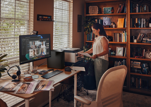Una mujer está en un escritorio usando una impresora mientras una pantalla de computadora muestra una videoconferencia.