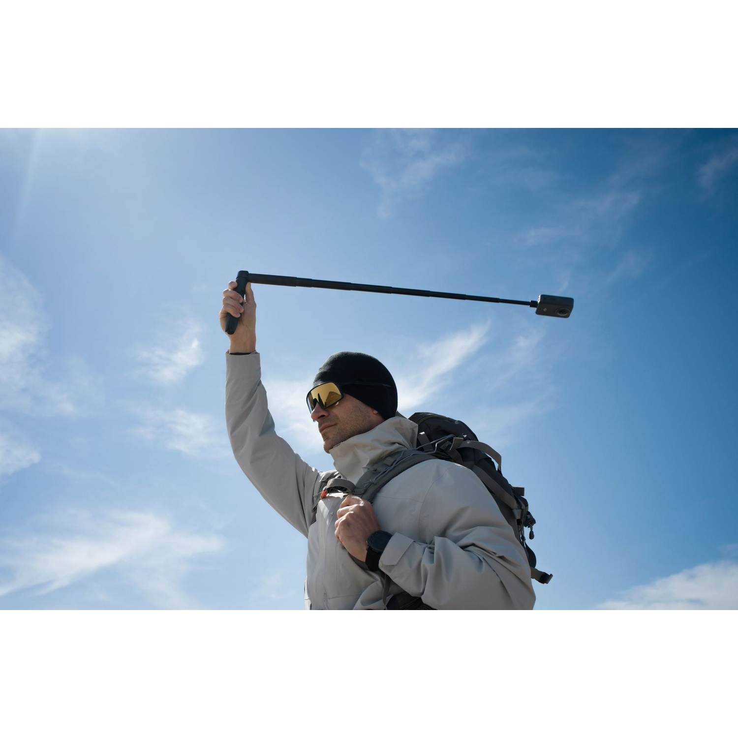Uomo con zaino tiene selfie stick contro cielo blu con nuvole.