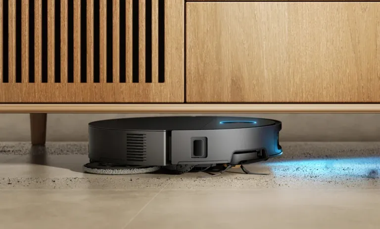 Un robot aspirador gris limpia debajo de un mueble de madera.