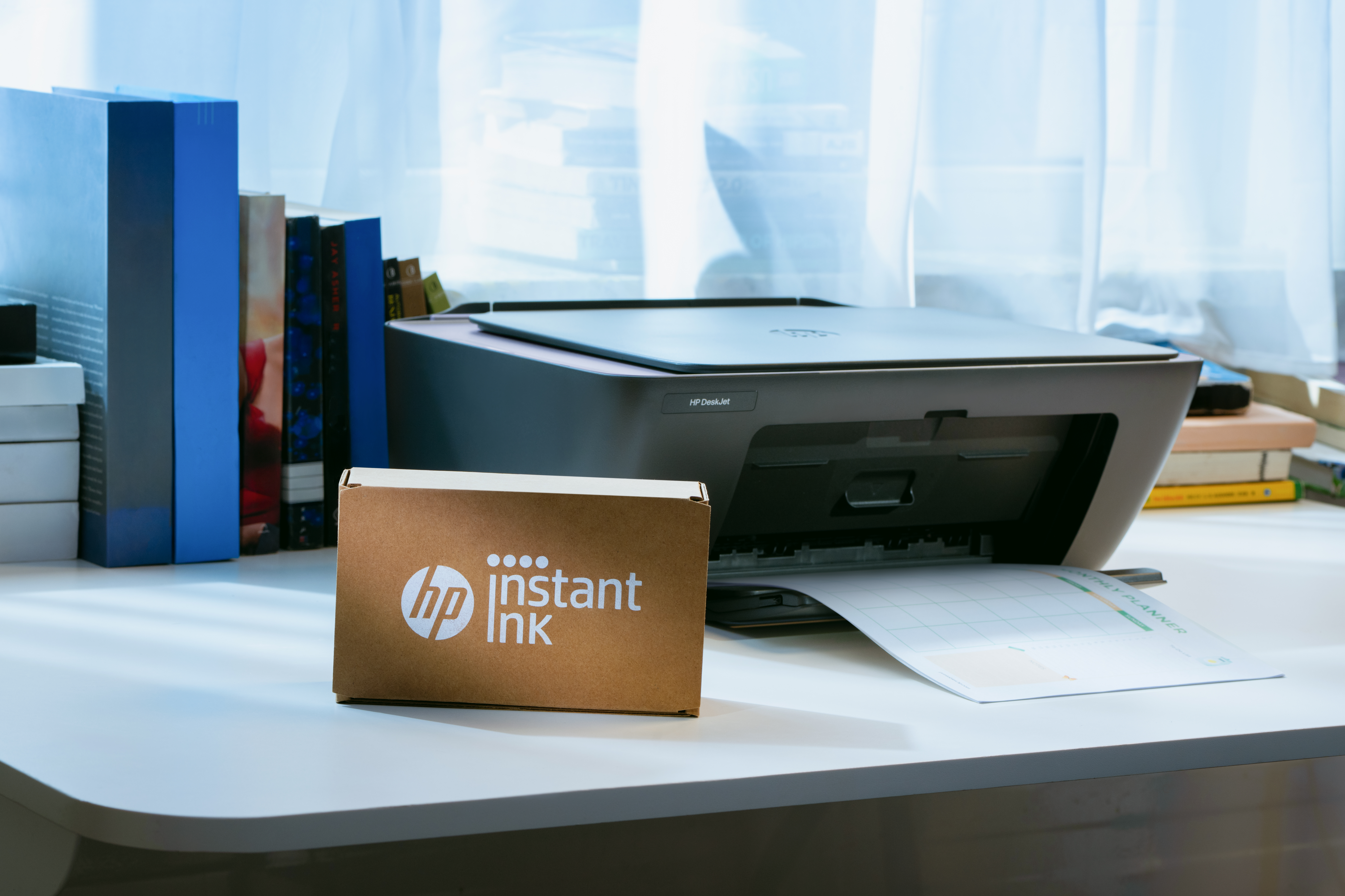 Ein HP Deskjet Drucker druckt einen Monatsplaner, daneben eine HP Instant Ink Box.