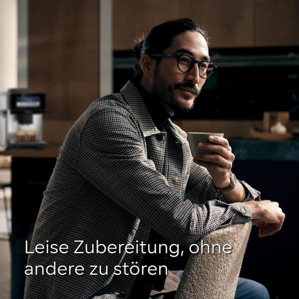 Mann mit Brille und Dutt hält Tasse, Kaffeemaschine im Hintergrund.