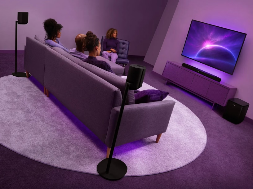 Emberek tévét néznek lila fényekkel és hangszórókkal megvilágított szobában.