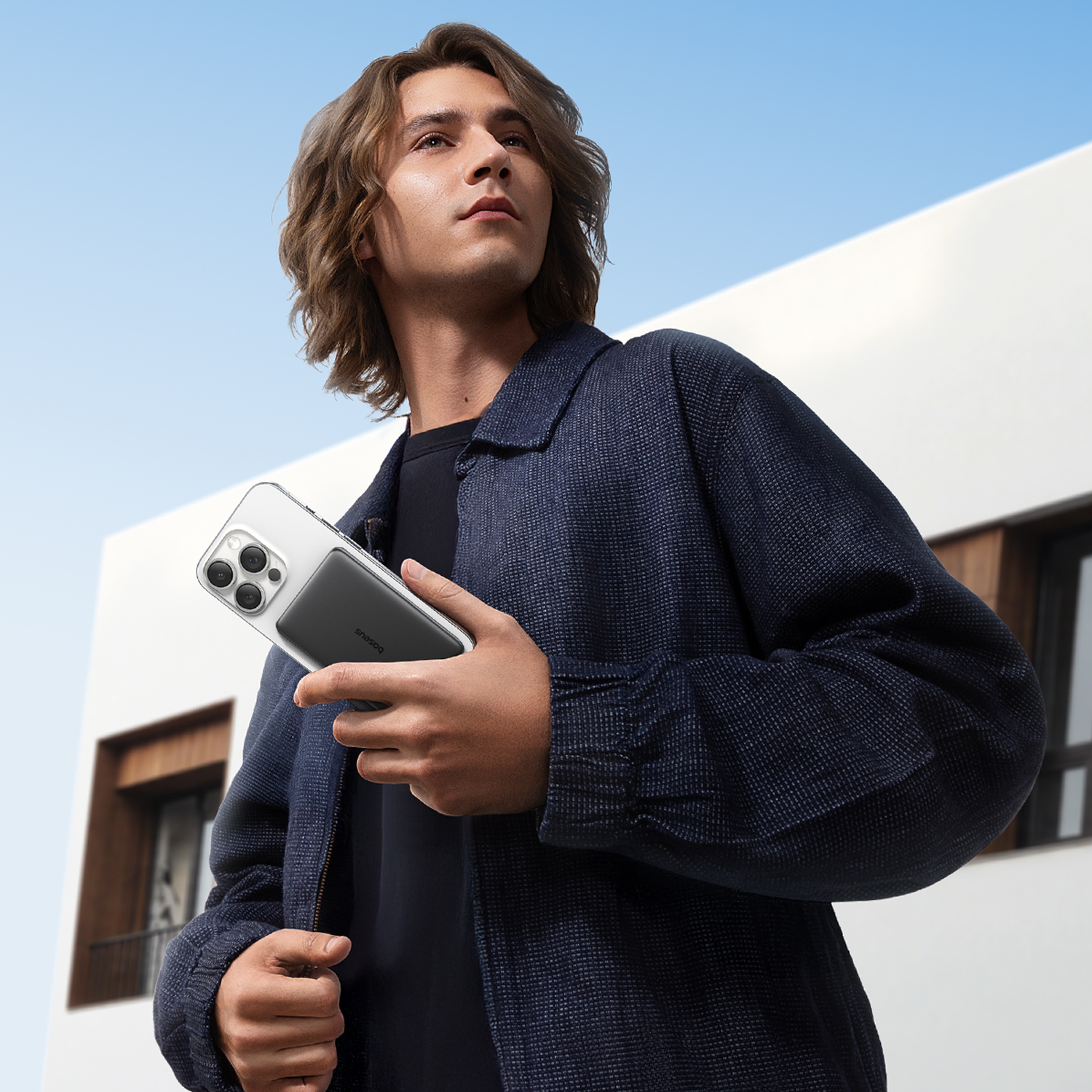 Un hombre de pelo largo sostiene un smartphone con funda frente a un edificio moderno.