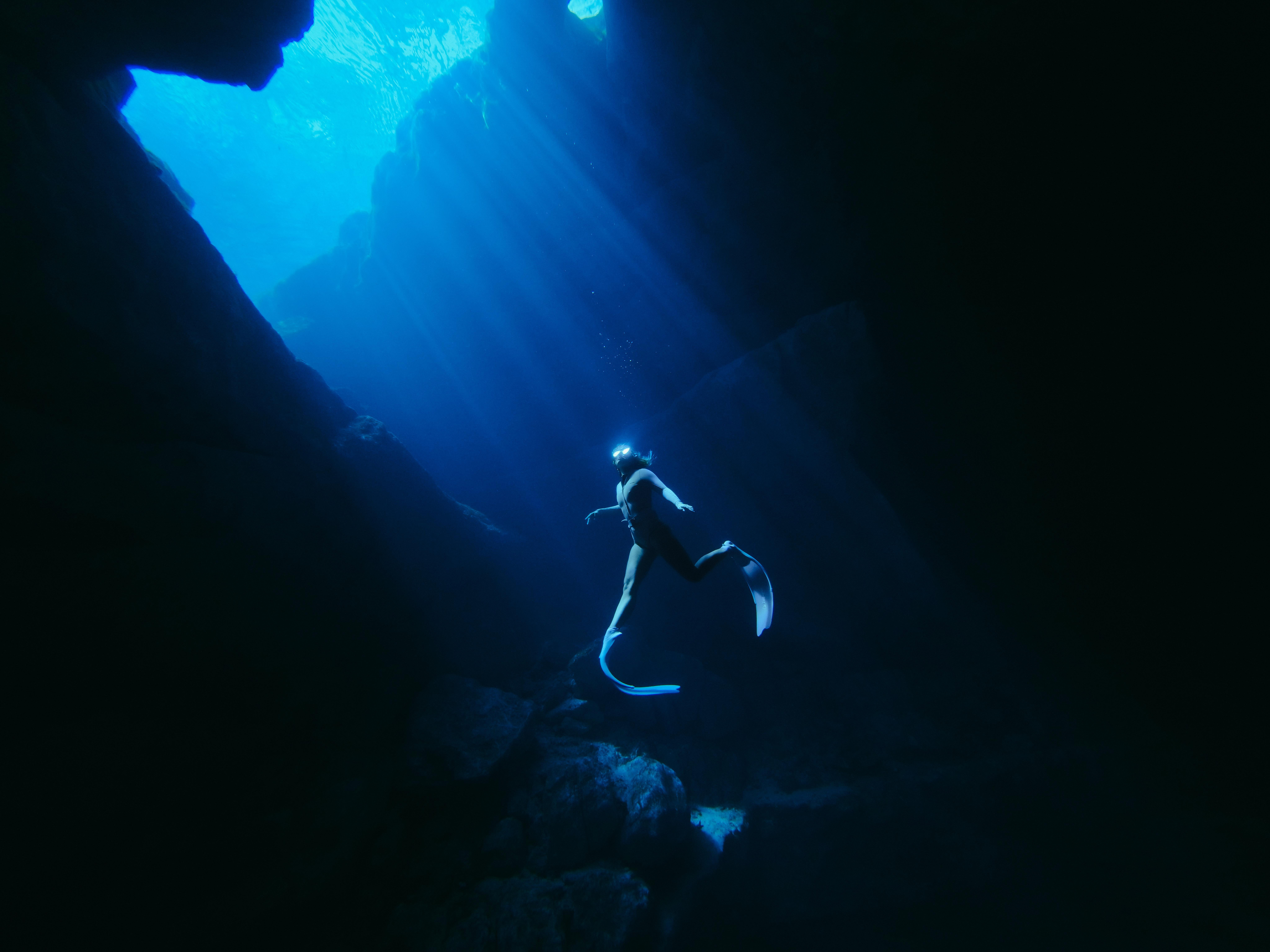 Ein Taucher mit Flossen schwimmt in einer dunklen Unterwasserhöhle, Sonnenstrahlen dringen ins Wasser.