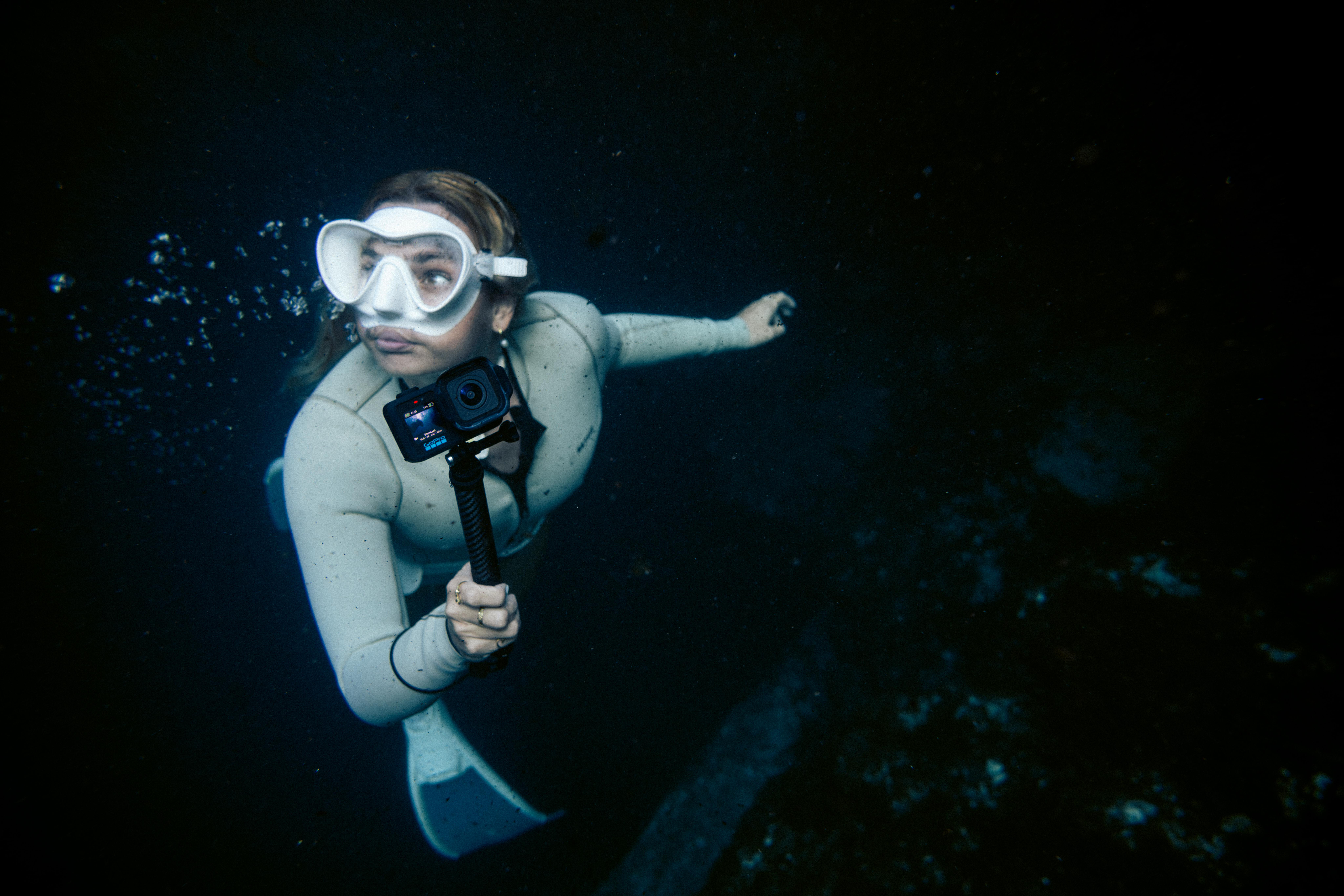 Eine Frau im hellen Neoprenanzug und mit Schnorchelmaske schwimmt unter Wasser und hält eine GoPro-Kamera.