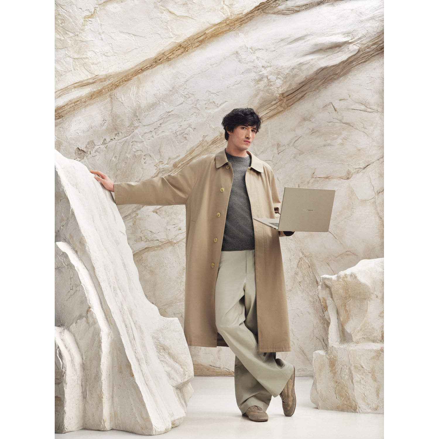 Un uomo con trench beige tiene un laptop in una stanza con pareti texturizzate, simili a rocce.