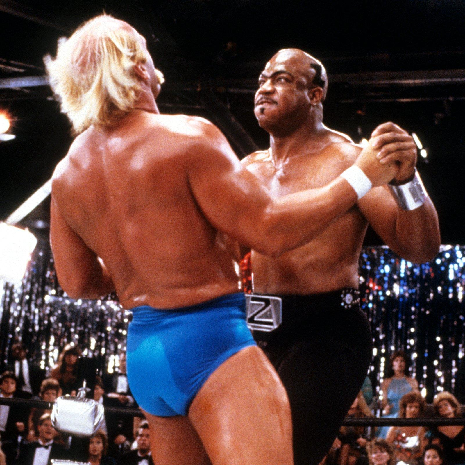 Zwei Wrestler im Ring, einer mit blonden Haaren, der andere mit Glatze. Glitzernder Hintergrund.