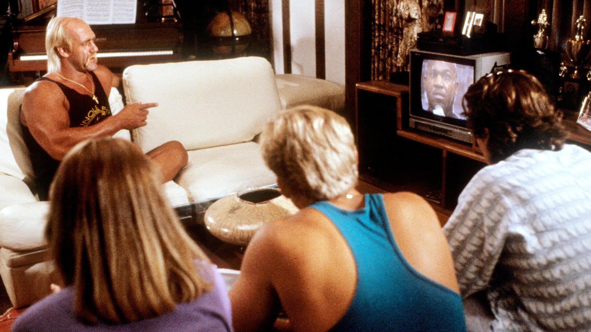 Vier Personen im Wohnzimmer schauen fern. Hulk Hogan zeigt auf den Bildschirm.
