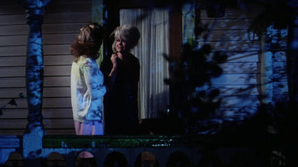 Zwei Frauen stehen auf einer Veranda bei Nacht. Eine trägt ein buntes Oberteil.