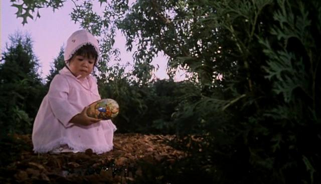 Ein Baby in rosa Haube und Kleid hält ein verziertes Ei in einem Waldgebiet.