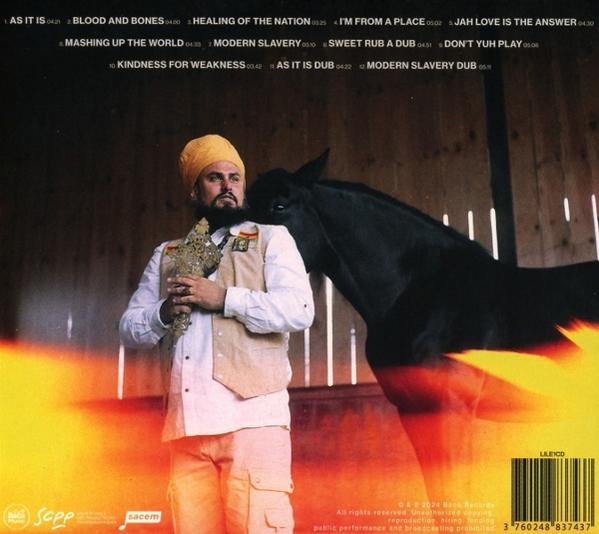 Mann mit gelbem Turban und Weste hält Kreuz vor schwarzem Pferd in Scheune.