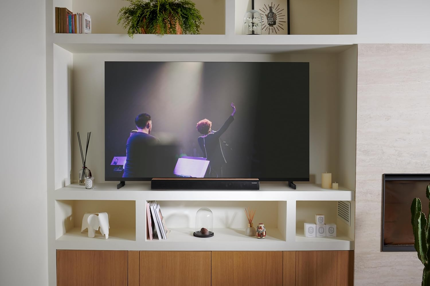 Un salón moderno con un televisor grande que muestra un concierto. Estantes con decoración, libros y plantas.