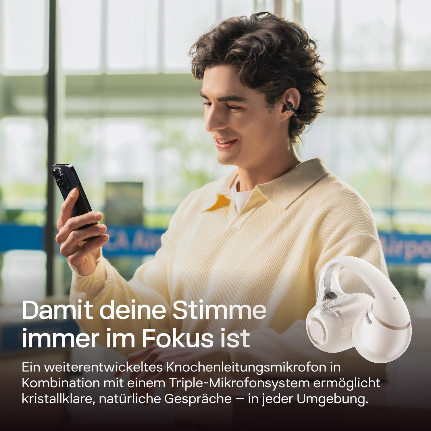 Ein Mann hält ein Smartphone und ein weißes Headset, im Hintergrund ein Flughafen.