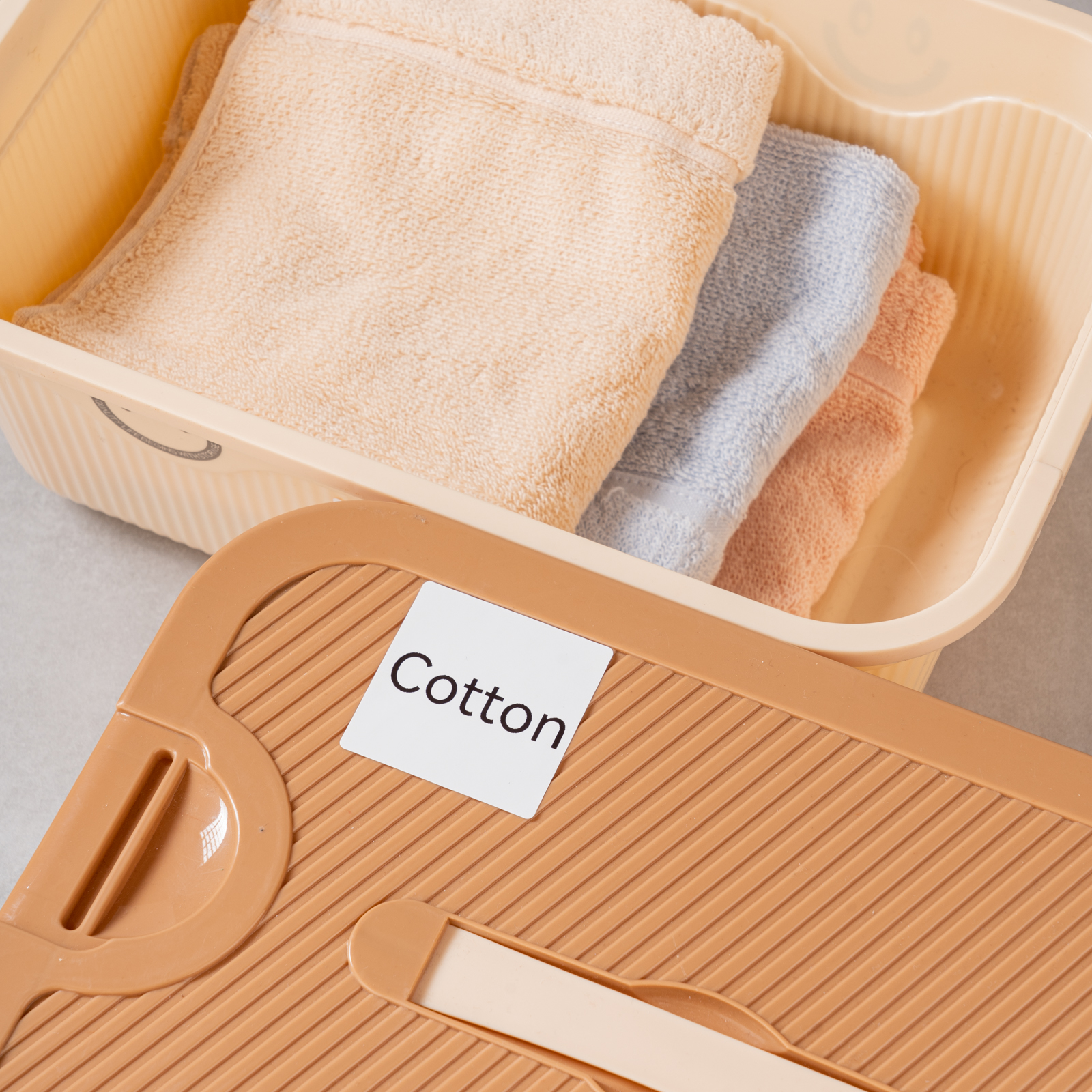 Ein beige Wanne mit gefalteten Handtüchern: hellbeige, hellblau und rosa. Ein braunes Brett mit Etikett 'Cotton'.