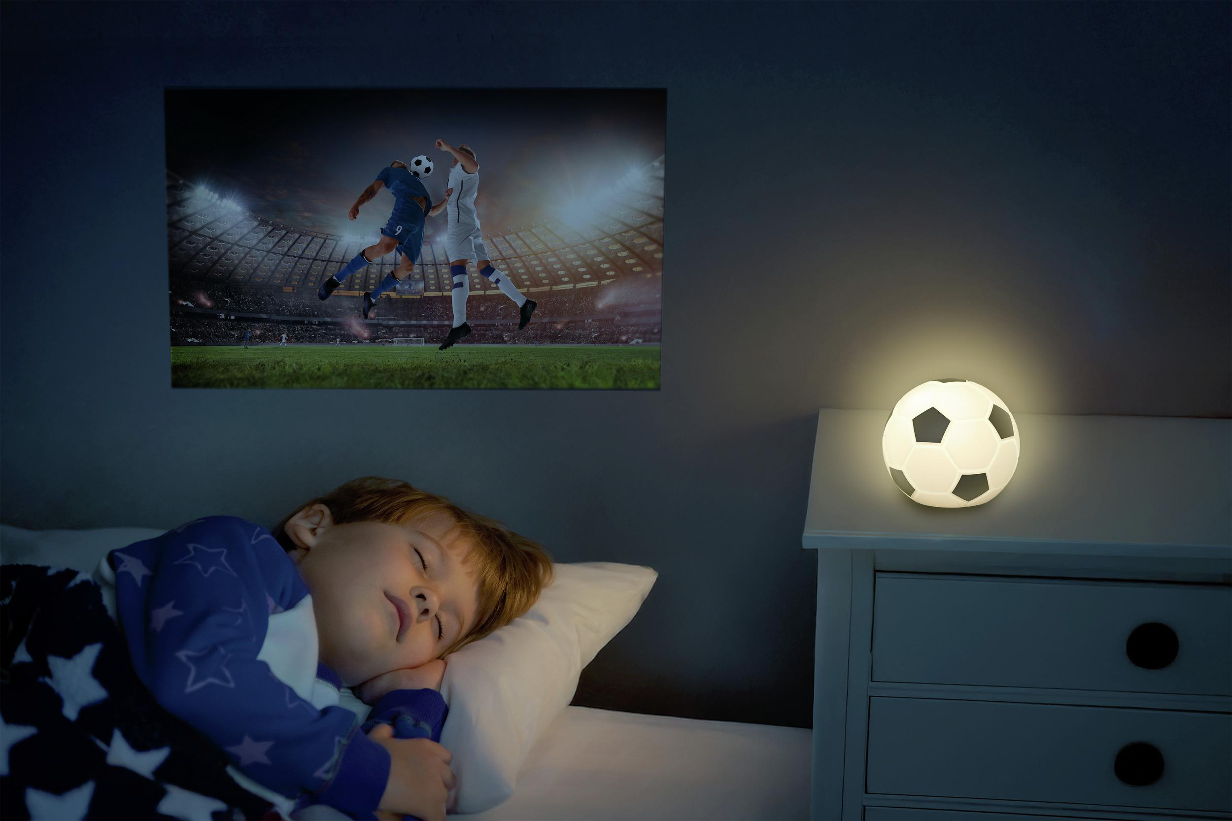 Ein Junge schläft im Bett, während ein Fußballspiel auf dem Bildschirm läuft. Ein Fußballlicht leuchtet.
