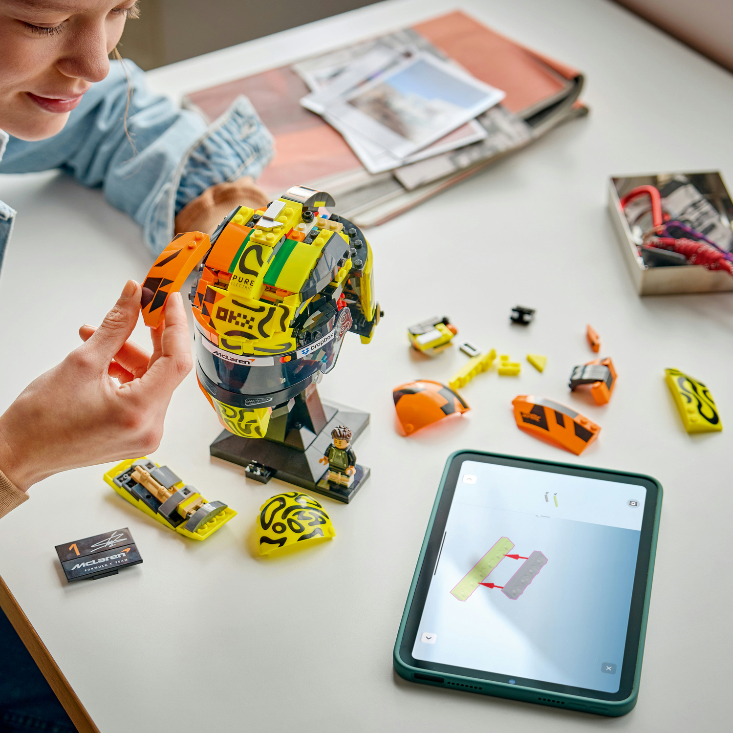 Una donna costruisce un casco Lego giallo con accenti arancioni. Pezzi sparsi intorno.