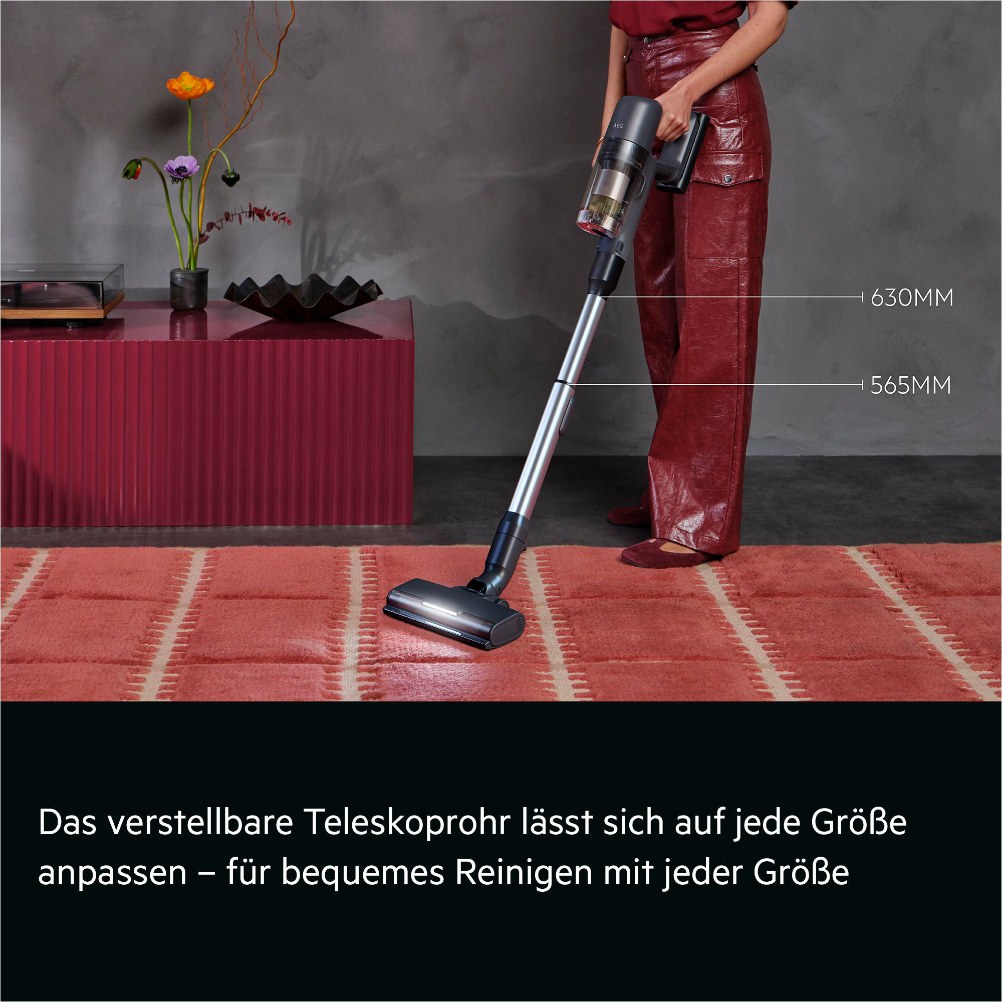Eine Person mit roten Hosen benutzt einen Akku-Staubsauger auf einem Teppich.