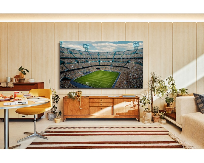 Telewizor wyświetla stadion piłkarski z kibicami. Stół jadalny i kanapa na pierwszym planie.